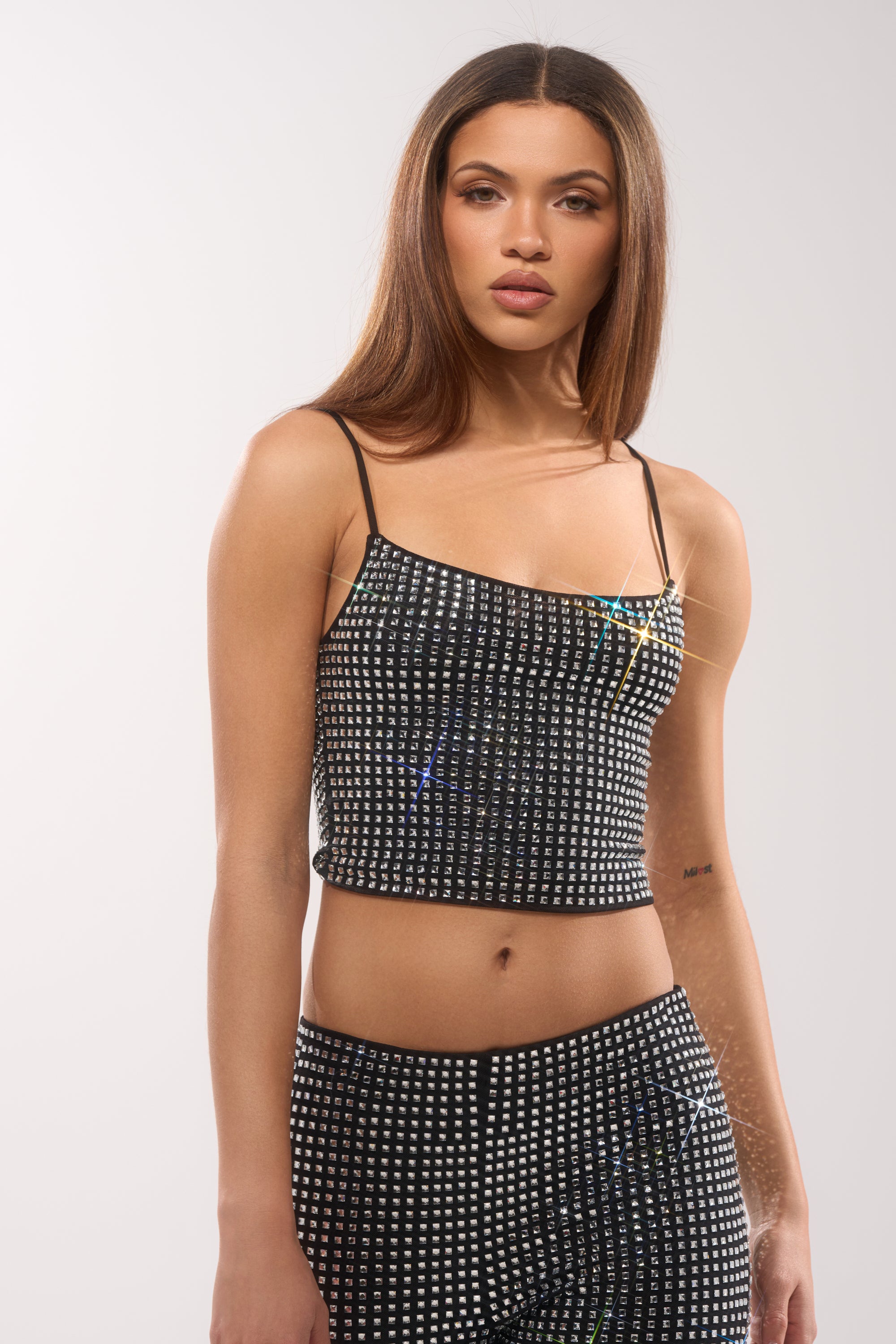 A woman with straight, long brown hair confidently poses against a plain light background, wearing the ALL EYES ON ME RHINESTONE BLOUSE—an eye-catching mesh crop top covered in small, shiny rhinestones. 1