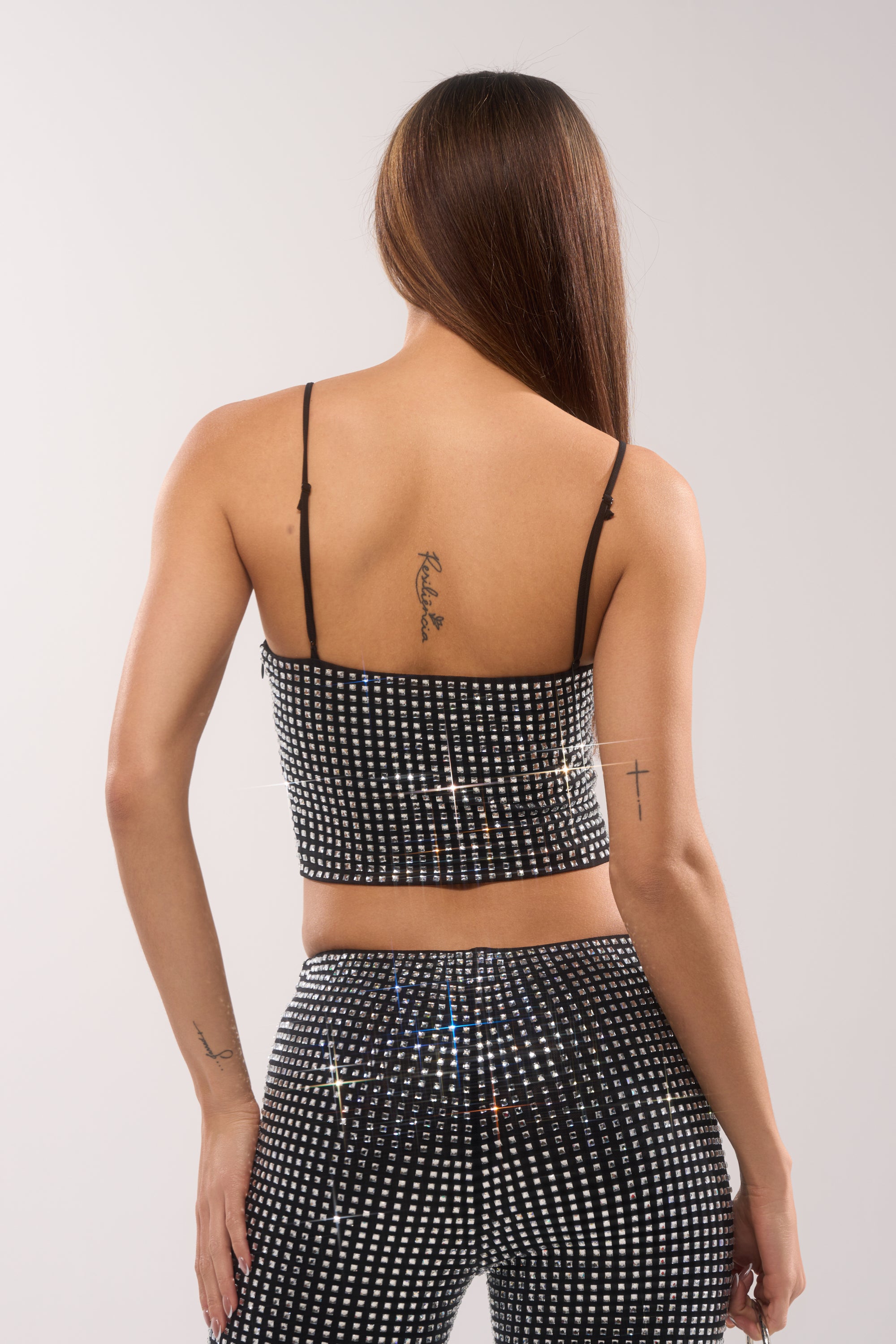 A woman with long brown hair, visible arm and shoulder blade tattoos, is seen from behind in a plain light setting wearing the ALL EYES ON ME RHINESTONE BLOUSE and matching pants in black and white. 4