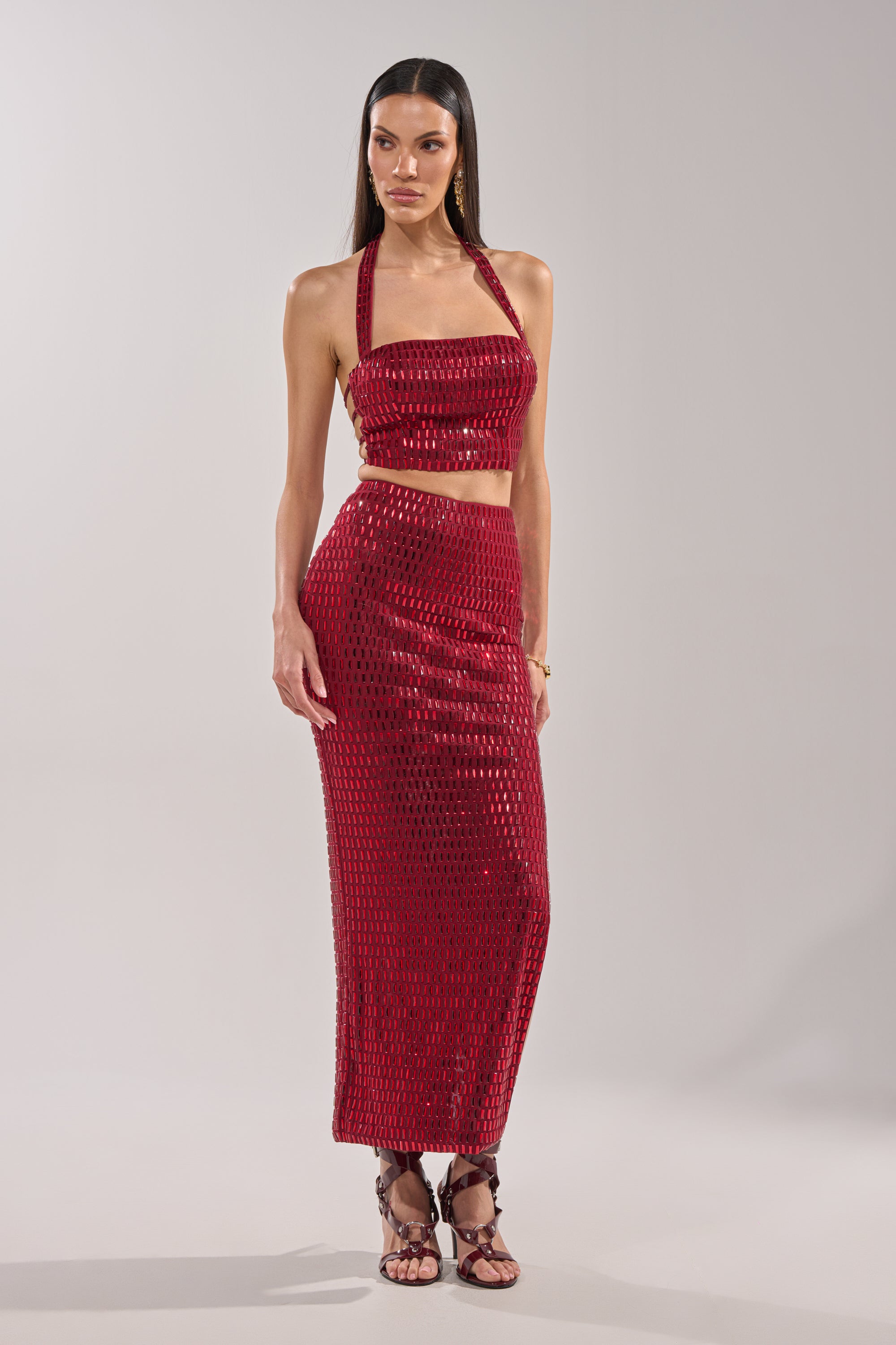 A woman wears the FALLING IN LOVE RHINESTONE MAXI SKIRT, a burgundy embellished maxi with a textured pattern, paired with a shiny red halter crop top and strappy sandals against a plain light background. 0