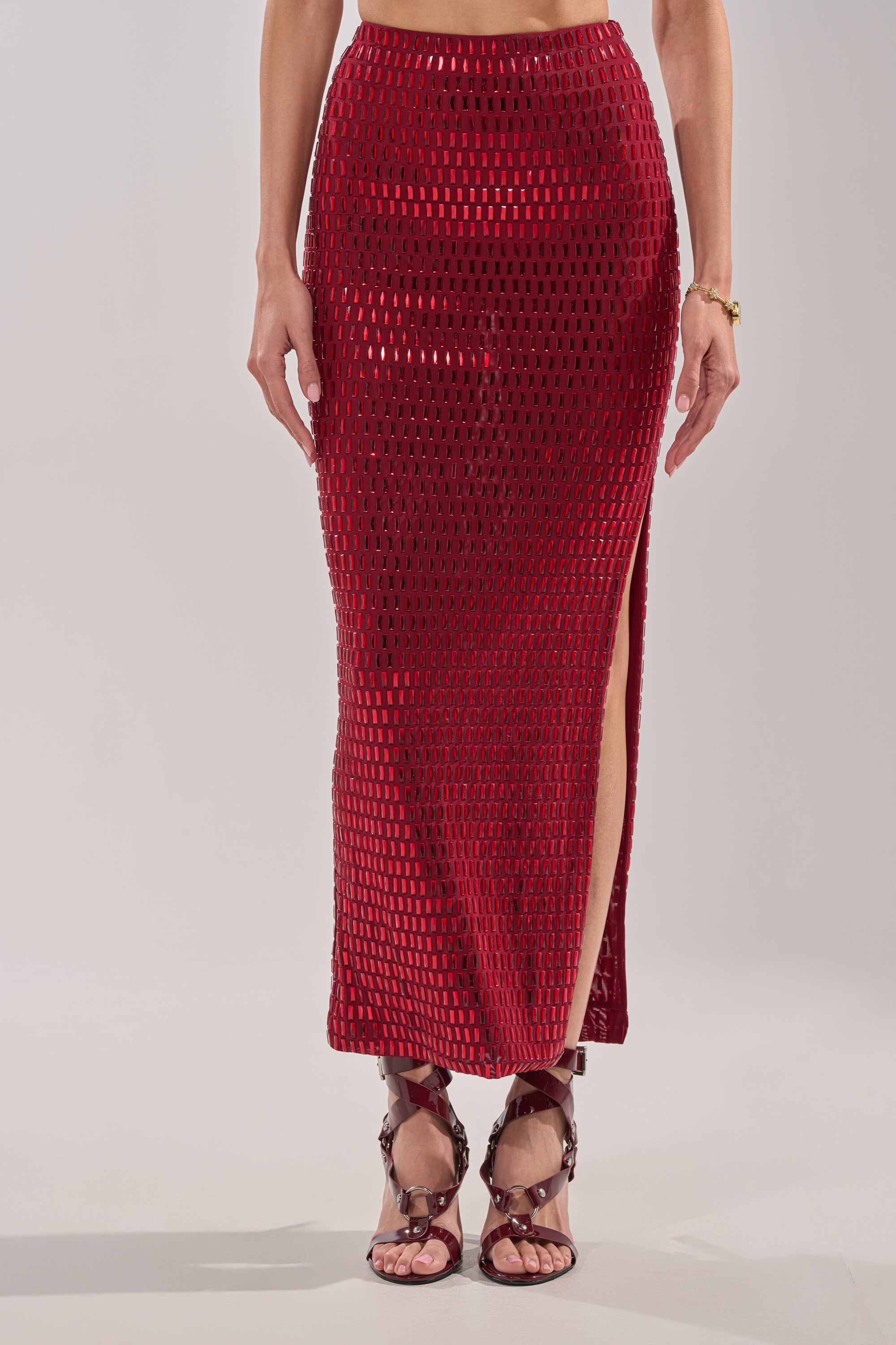 Someone wears the FALLING IN LOVE RHINESTONE MAXI SKIRT in burgundy, featuring a textured square pattern, rhinestone embellishments, and a high slit. It's styled with strappy brown heels and a simple bracelet against a plain light background. 1