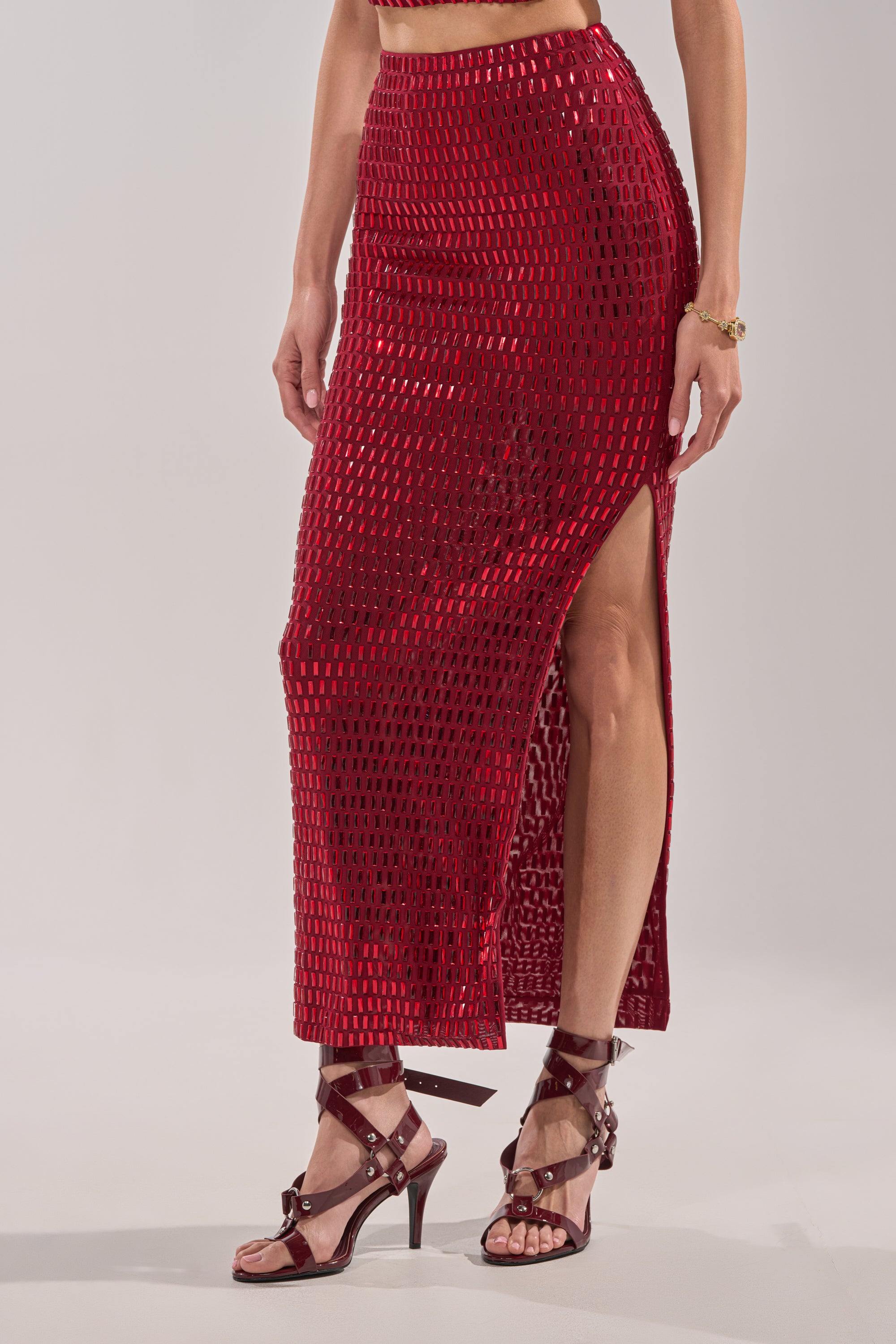 A woman wears the FALLING IN LOVE RHINESTONE MAXI SKIRT, a shiny red piece with a side slit and geometric cutouts, paired with strappy red high-heeled sandals. Only the lower half of her body is visible. 2
