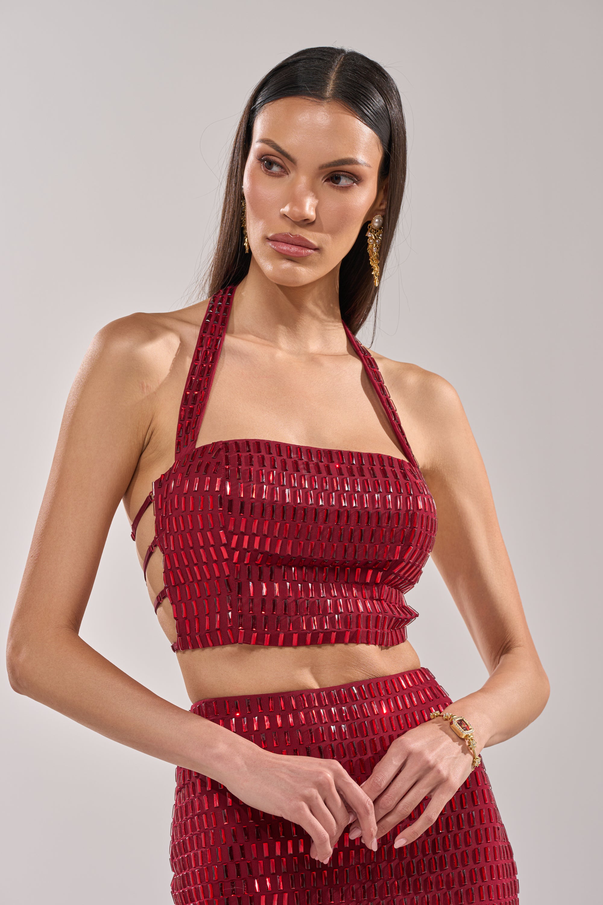 A woman with long straight hair models the FALLING IN LOVE RHINESTONE BLOUSE, featuring mesh fabric and side strap details, paired with a matching skirt. She completes the look with gold earrings and a bracelet against a plain light background. 0