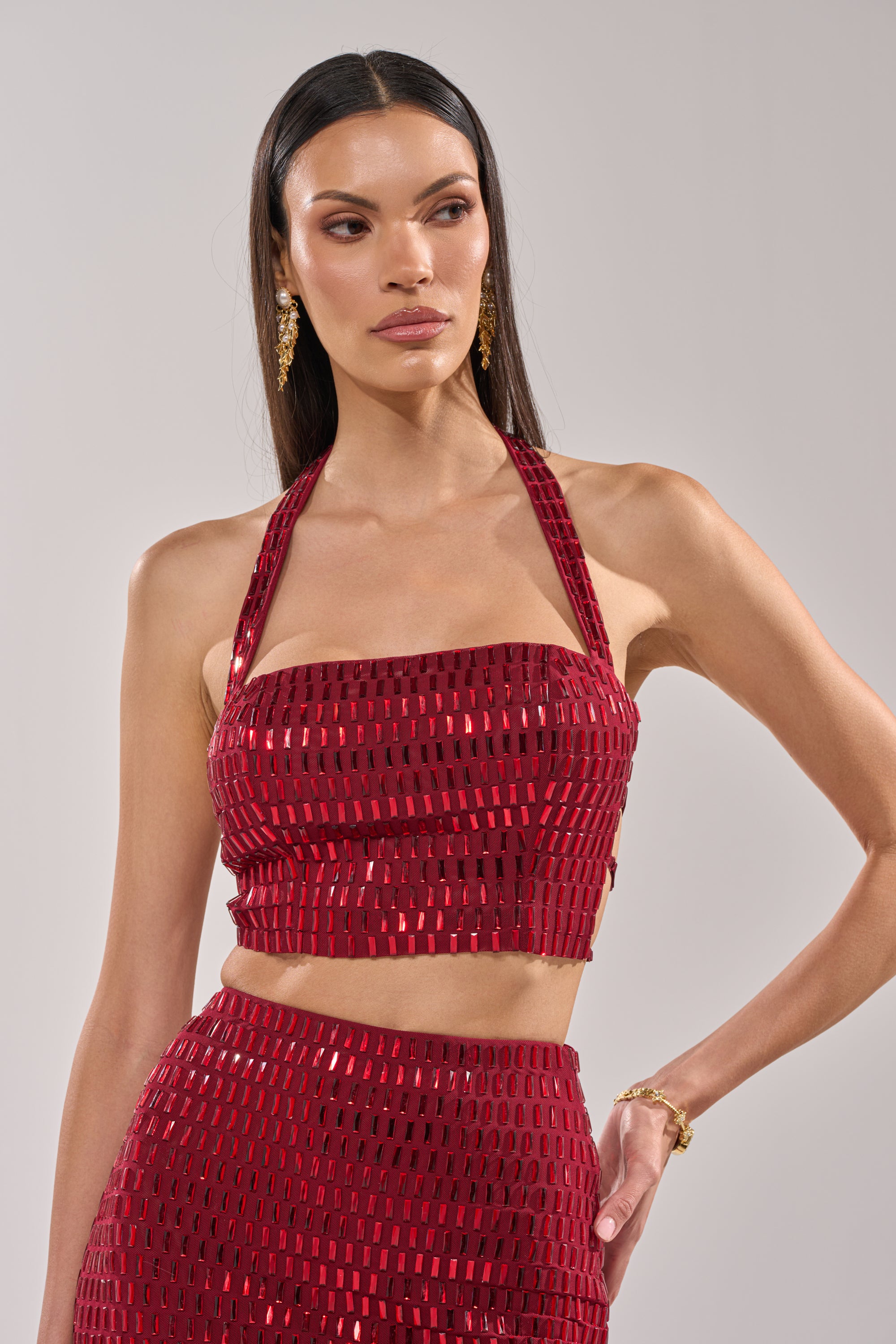 A woman wearing the FALLING IN LOVE RHINESTONE BLOUSE, a burgundy embellished halter top with adjustable elastic back straps. 1