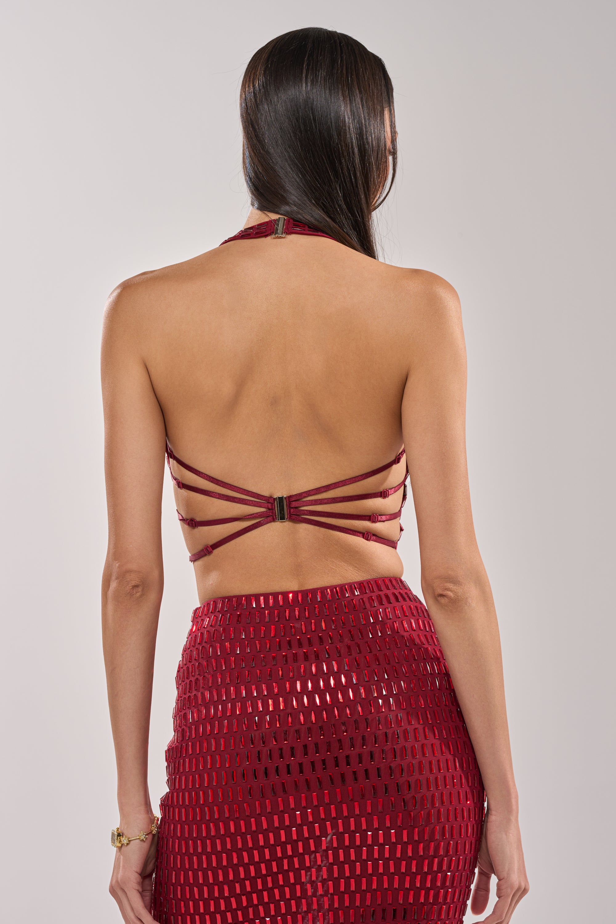 A woman with long dark hair is seen from the back, wearing the FALLING IN LOVE RHINESTONE BLOUSE in burgundy with adjustable back straps and embellishments, paired with a matching red geometric patterned skirt. 4