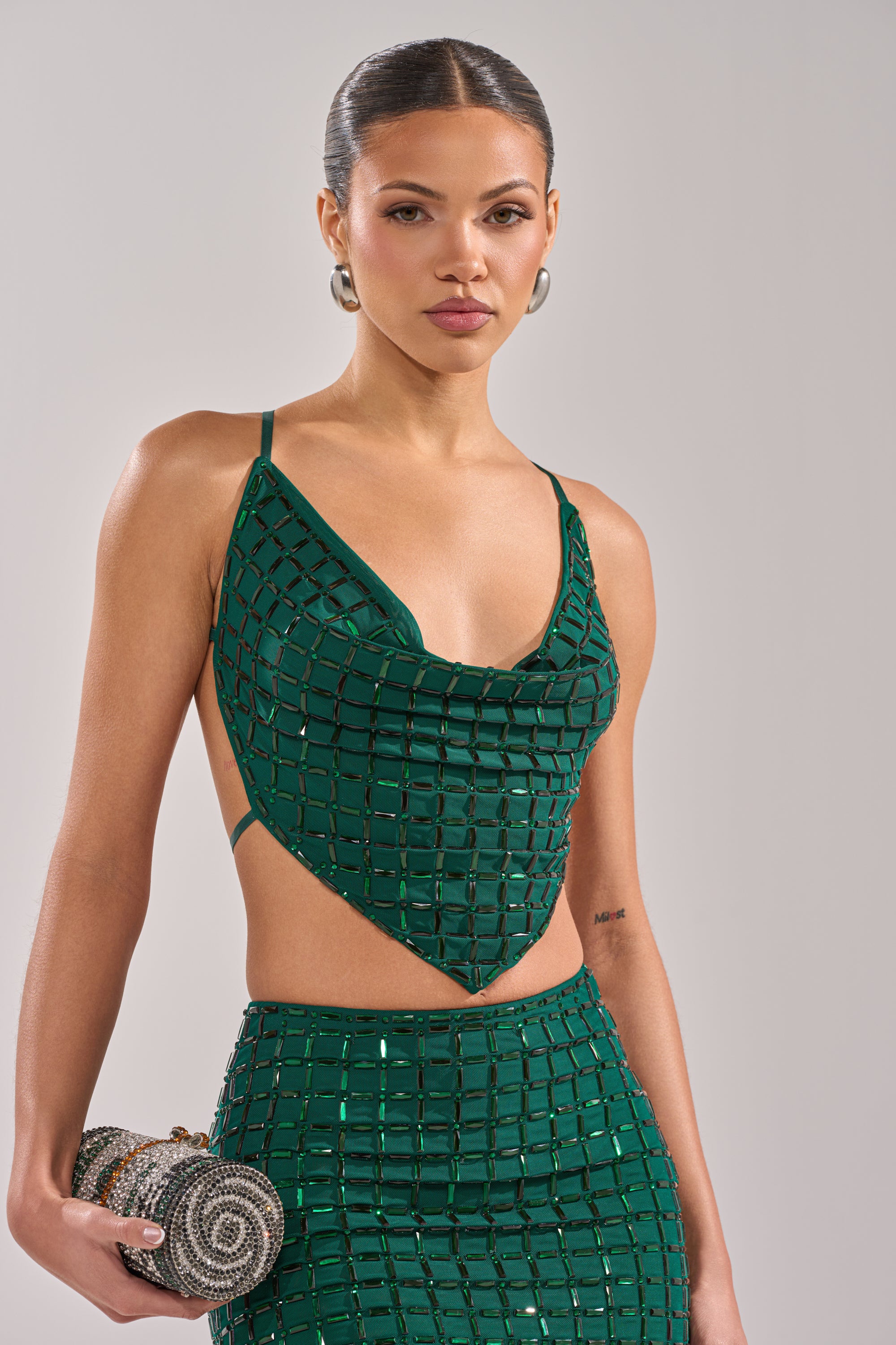 A woman with slicked-back hair wears the PICK ME A WINNER RHINESTONE TANK, an emerald green, grid-patterned sequined crop top, paired with a matching skirt and hoop earrings. She holds a round clutch and poses confidently against a neutral background. 0