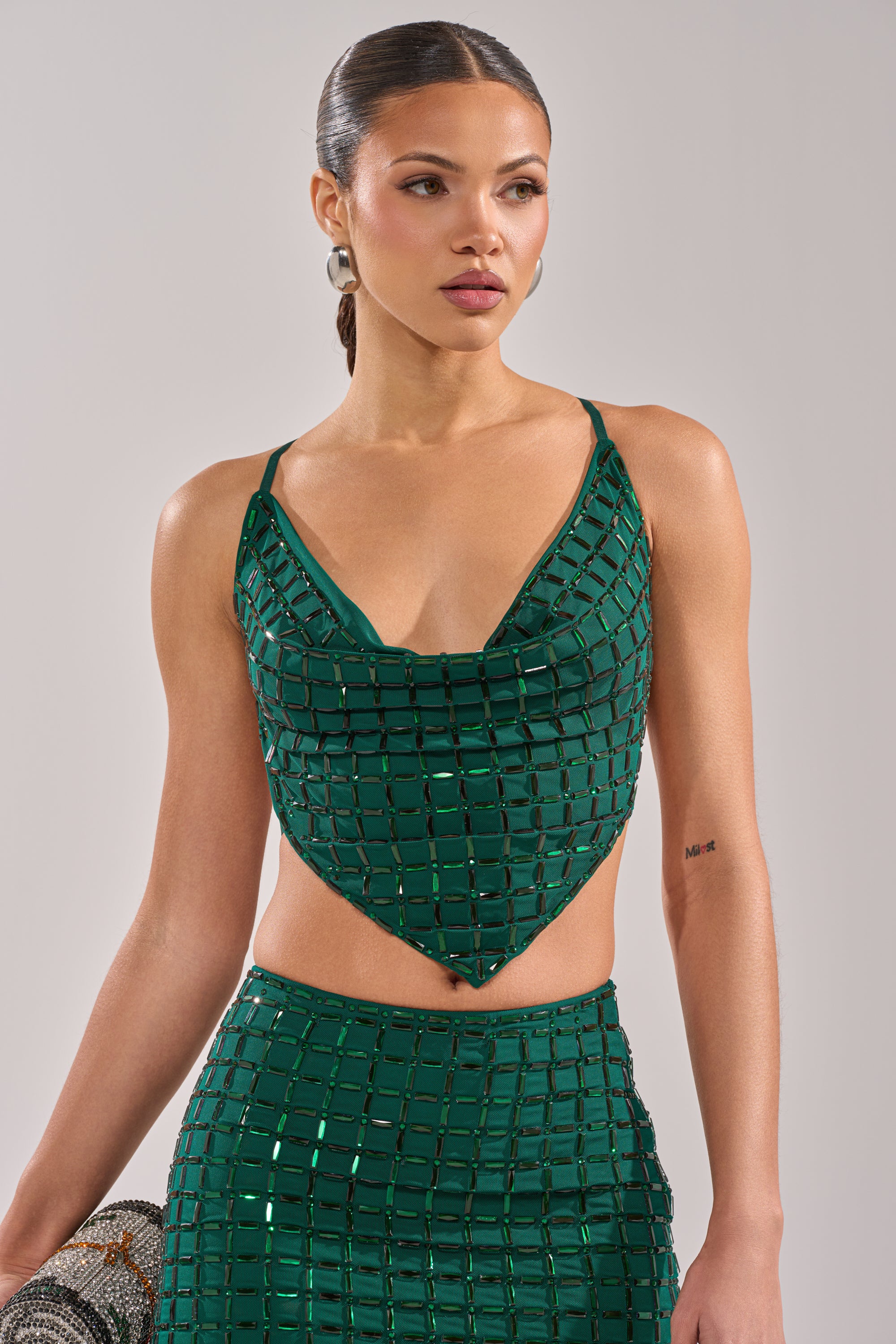 A woman wears the PICK ME A WINNER RHINESTONE TANK—an emerald green checkered crop top with thin straps and rhinestone accents—paired with a matching skirt and large hoop earrings, posing against a plain background. 1