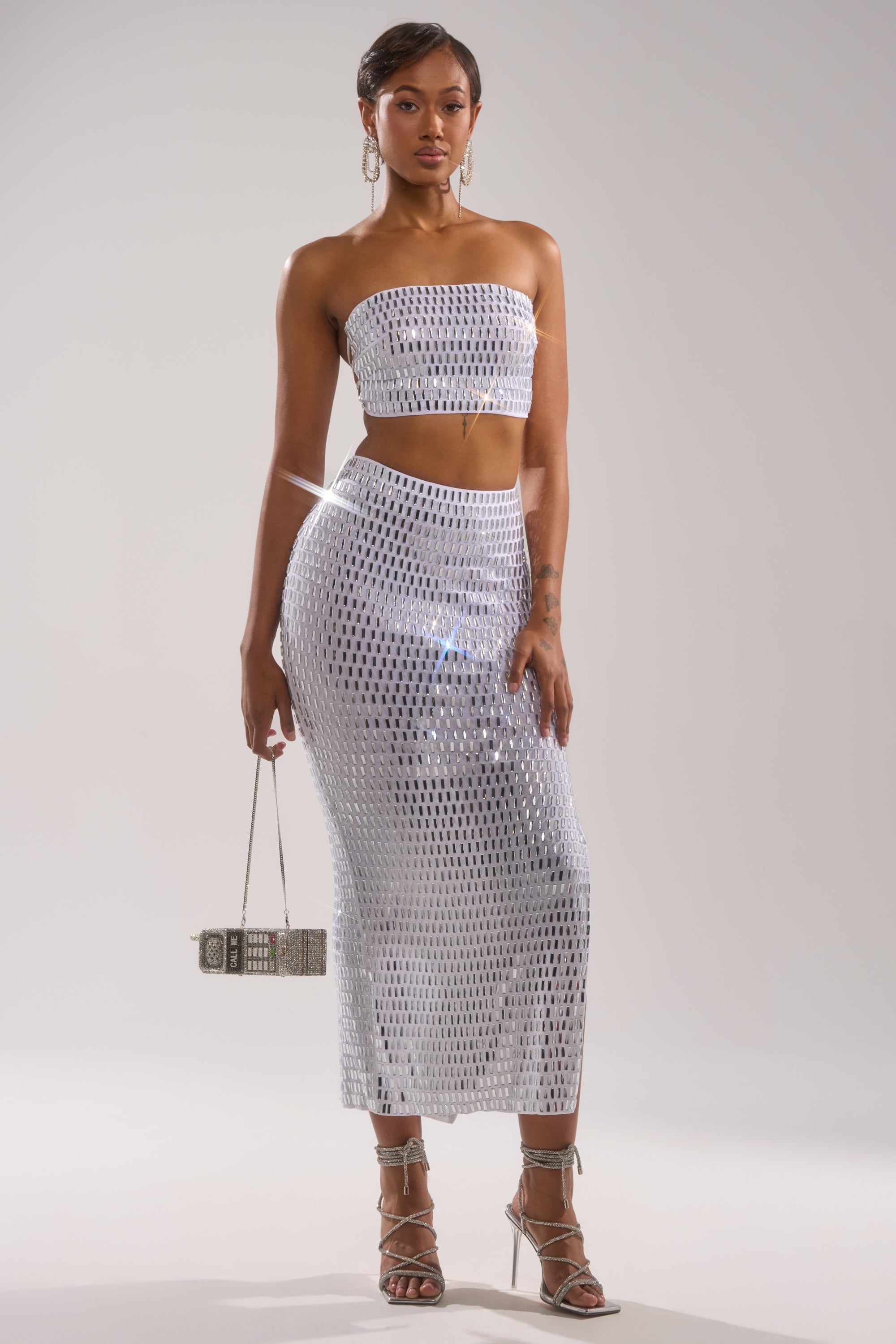 A woman poses in a strapless silver sequin crop top and TALK ABOUT ME RHINESTONE MAXI SKIRT, holding a small silver handbag and wearing strappy silver heels against a plain light background.