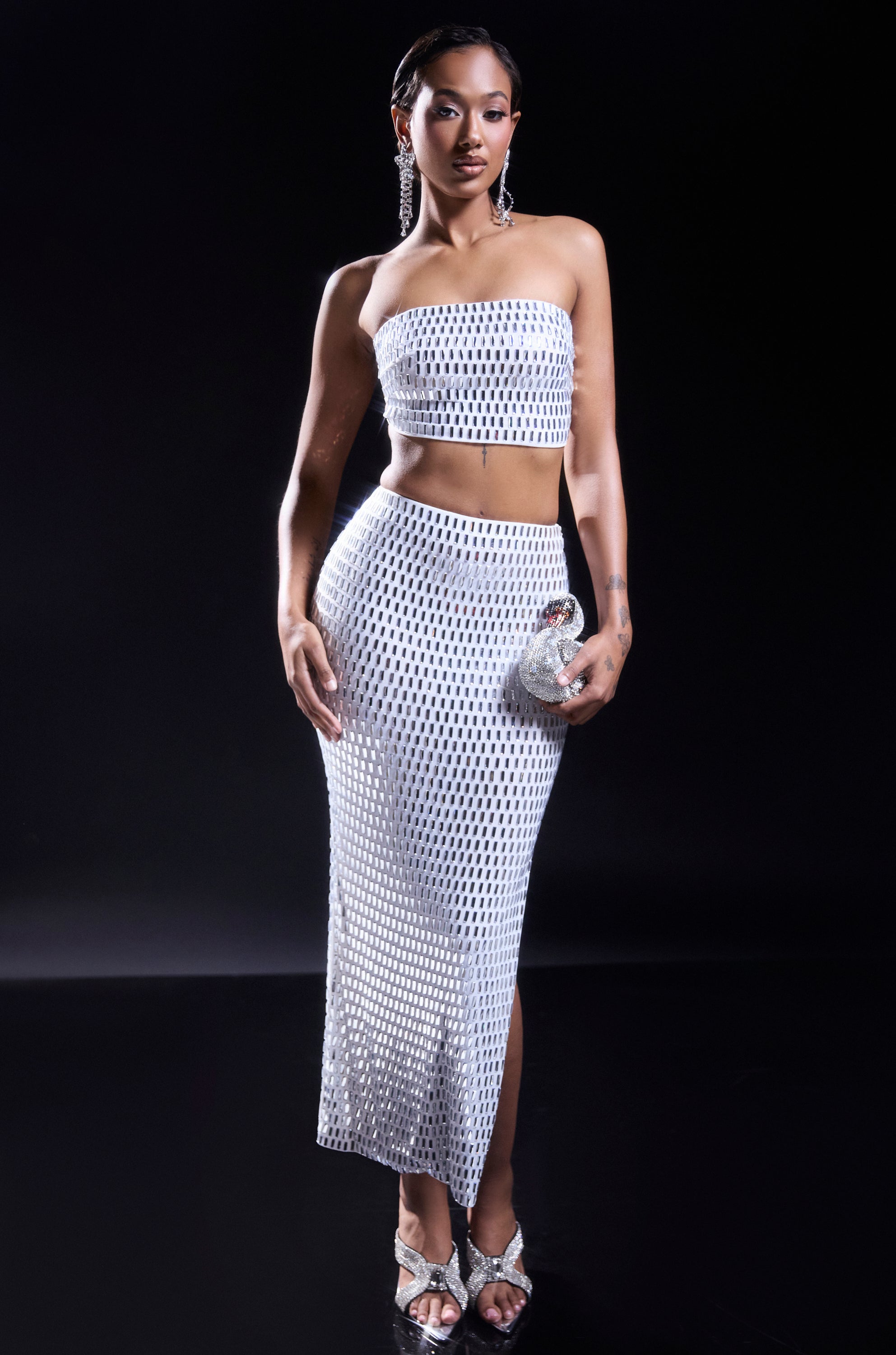 A woman stands against a black background in an AKIRA Label white, strapless crop top and the TALK ABOUT ME RHINESTONE MAXI SKIRT. She accessorizes with a small clutch, silver heels, statement earrings, and sleek straight hair.