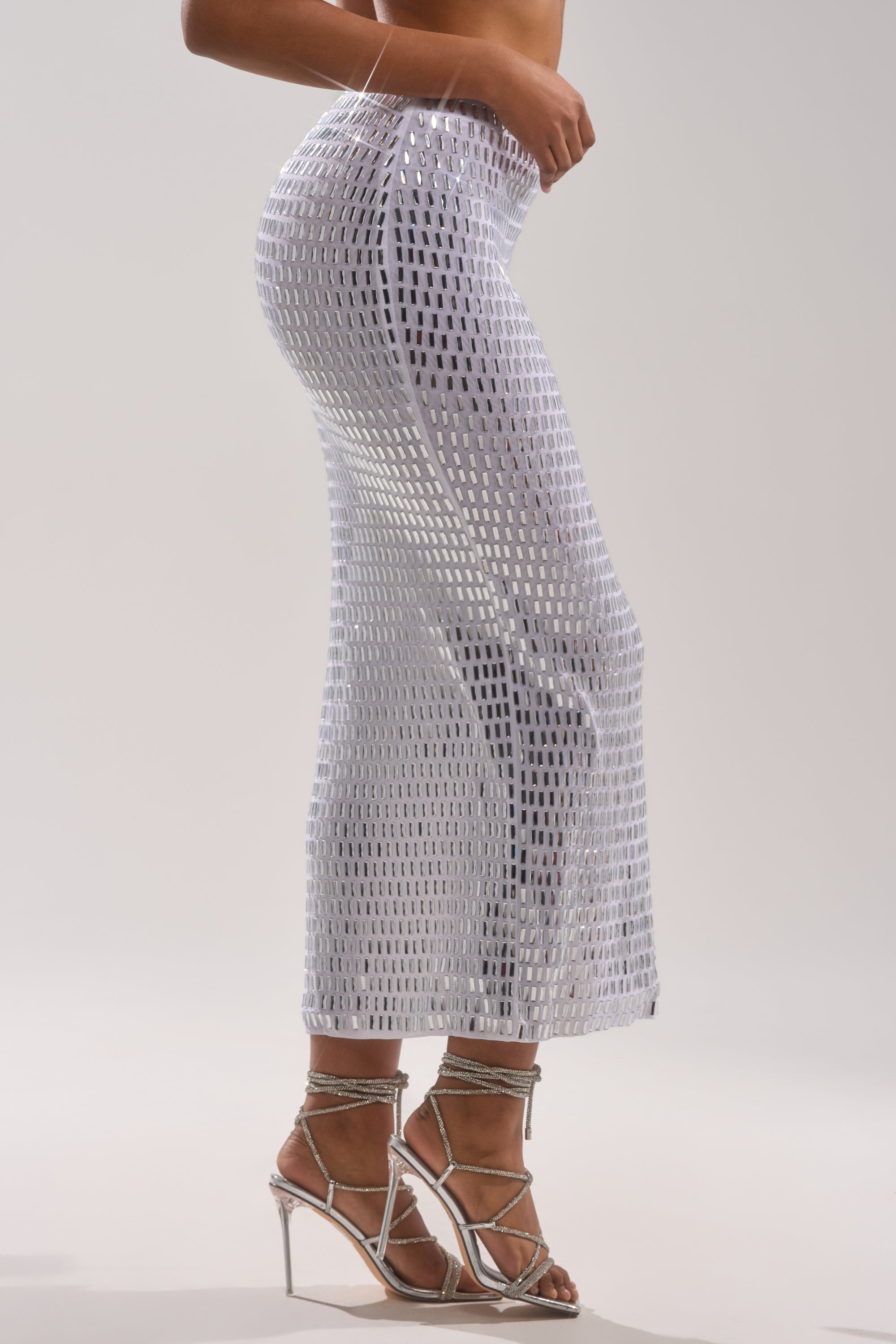 A person shown from the side against a plain light background wears the TALK ABOUT ME RHINESTONE MAXI SKIRT in silver metallic mesh with strappy silver high-heeled sandals.