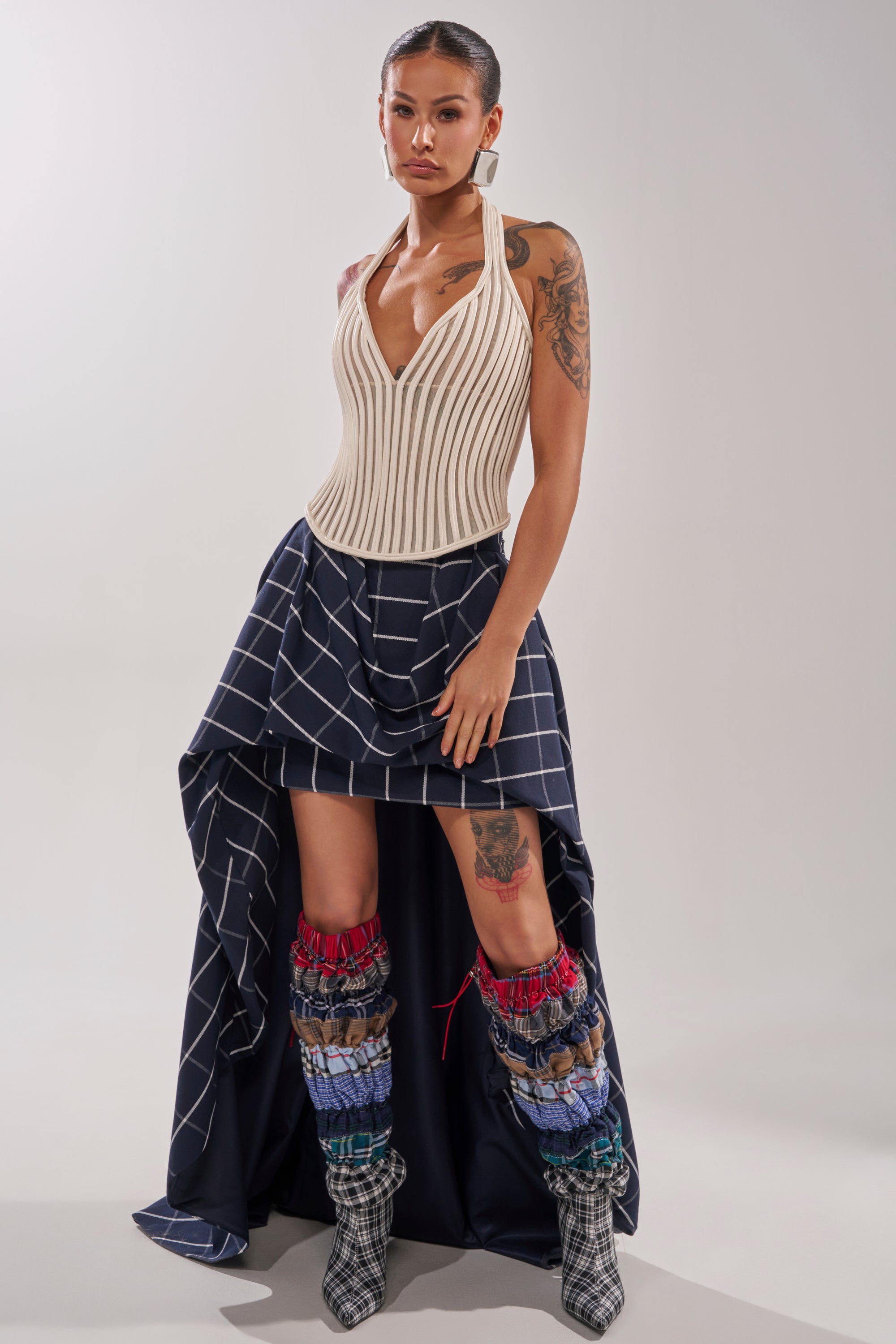 A woman stands confidently in the MAKE A STATEMENT HIGH LOW SKIRT, featuring an asymmetrical plaid design with white grid lines, paired with a ribbed cream top, colorful leg warmers, checkered boots, and visible tattoos. Her hair is pulled back. 1