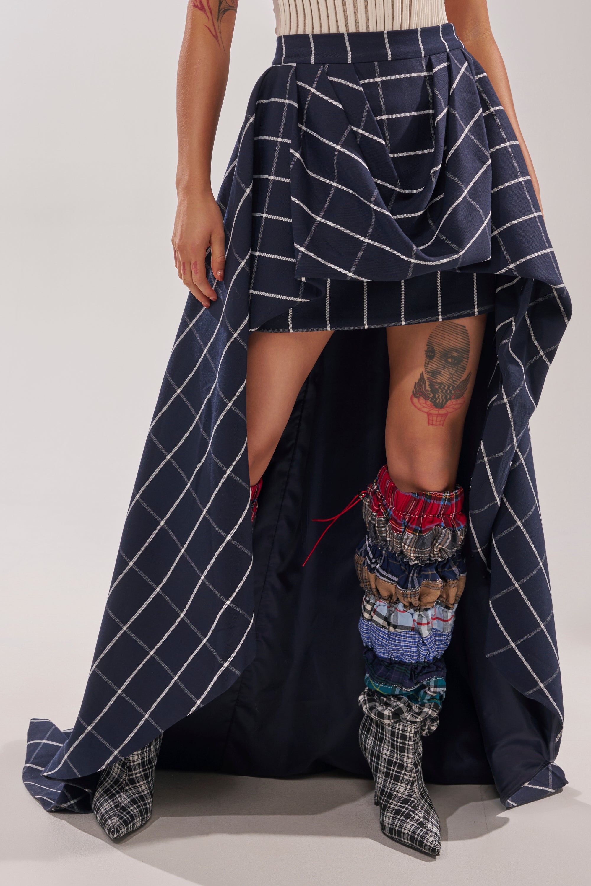 A person wearing the MAKE A STATEMENT HIGH LOW SKIRT in navy blue and white plaid with a high slit, pairing it with textured multi-colored leg warmers and pointed-toe plaid boots, revealing a skull tattoo on one leg. 0