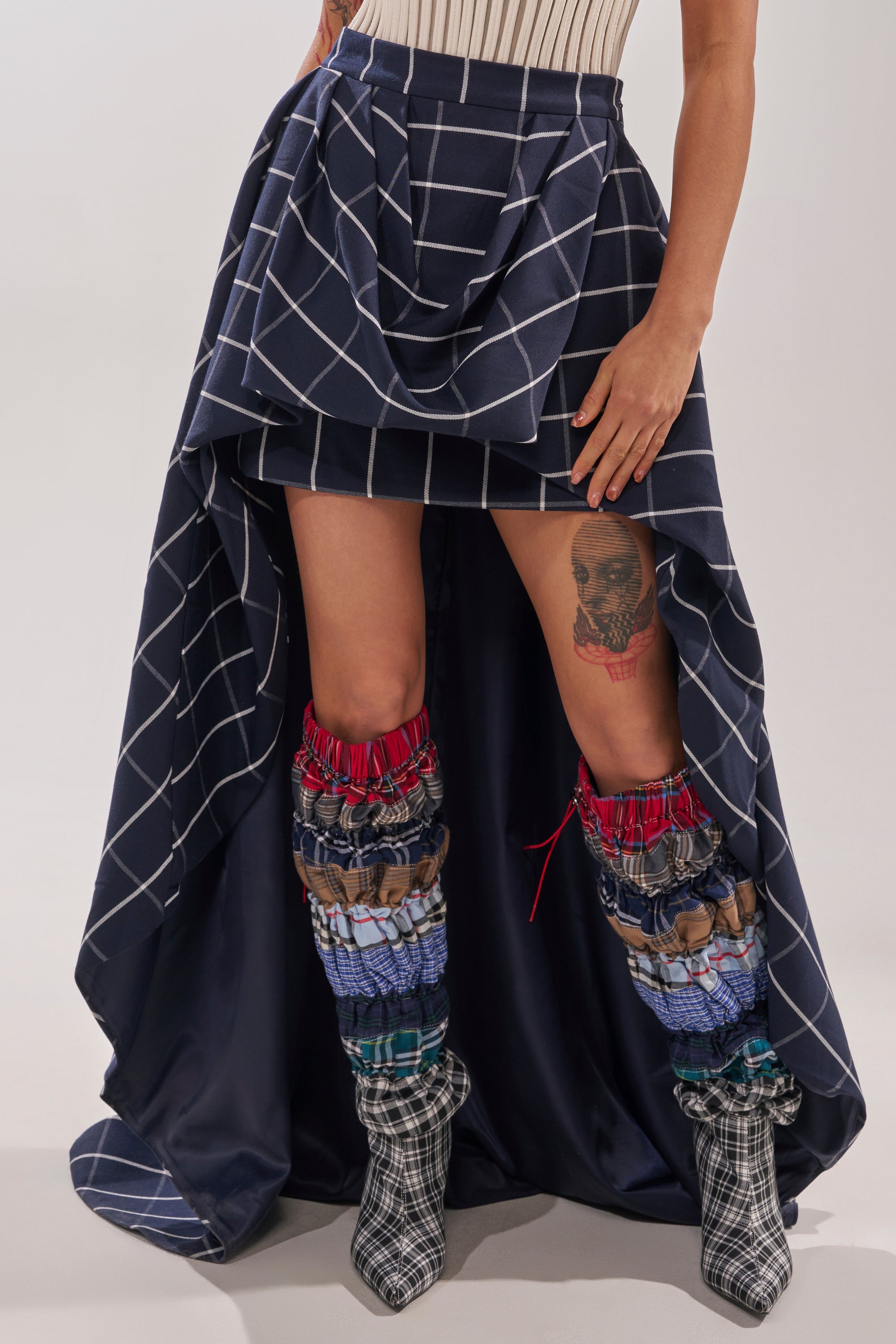 A person wears the MAKE A STATEMENT HIGH LOW SKIRT in navy blue with white grid lines, paired with a beige ribbed top and multicolored patchwork boots, showcasing a thigh tattoo while their hands rest on the statement skirt. 2