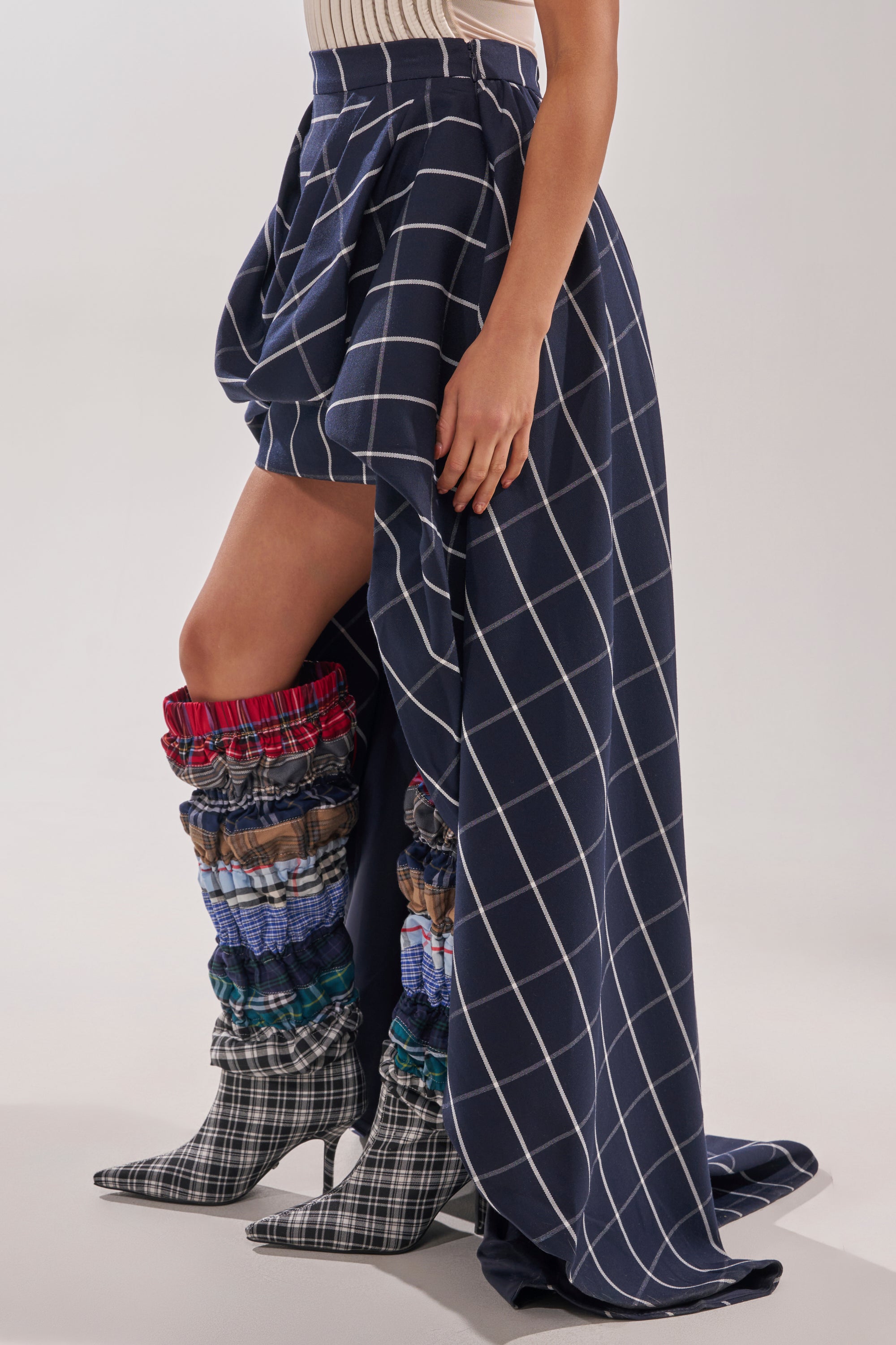 Wearing the MAKE A STATEMENT HIGH LOW SKIRT in blue with white grid lines, a person reveals multi-patterned ruched boots with pointed toes and plaid details while standing against a plain background. 3