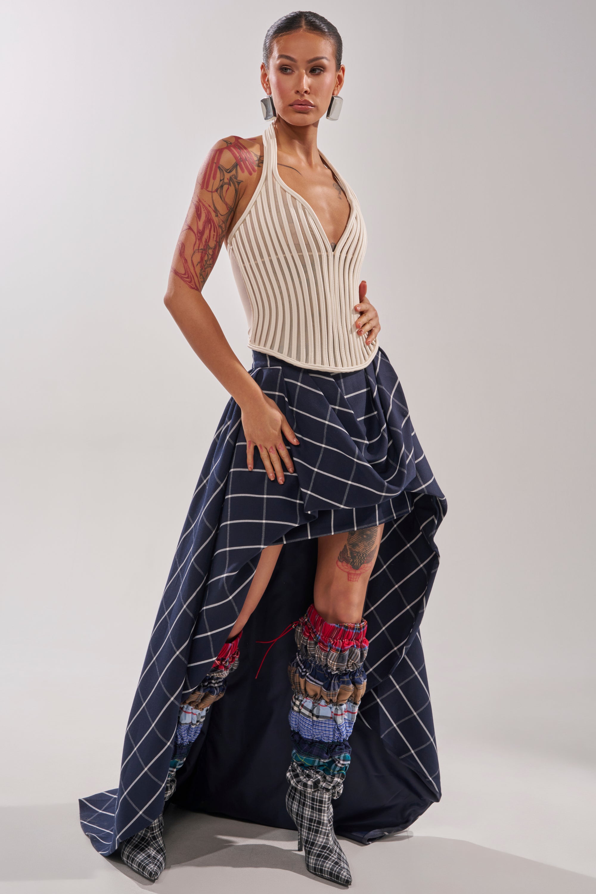 A woman showcases the MAKE A STATEMENT HIGH LOW SKIRT with a cream halter top and layered multicolored boots, accessorizing with large earrings. Her visible arm tattoos stand out against the plain background for a bold, modern look. 6