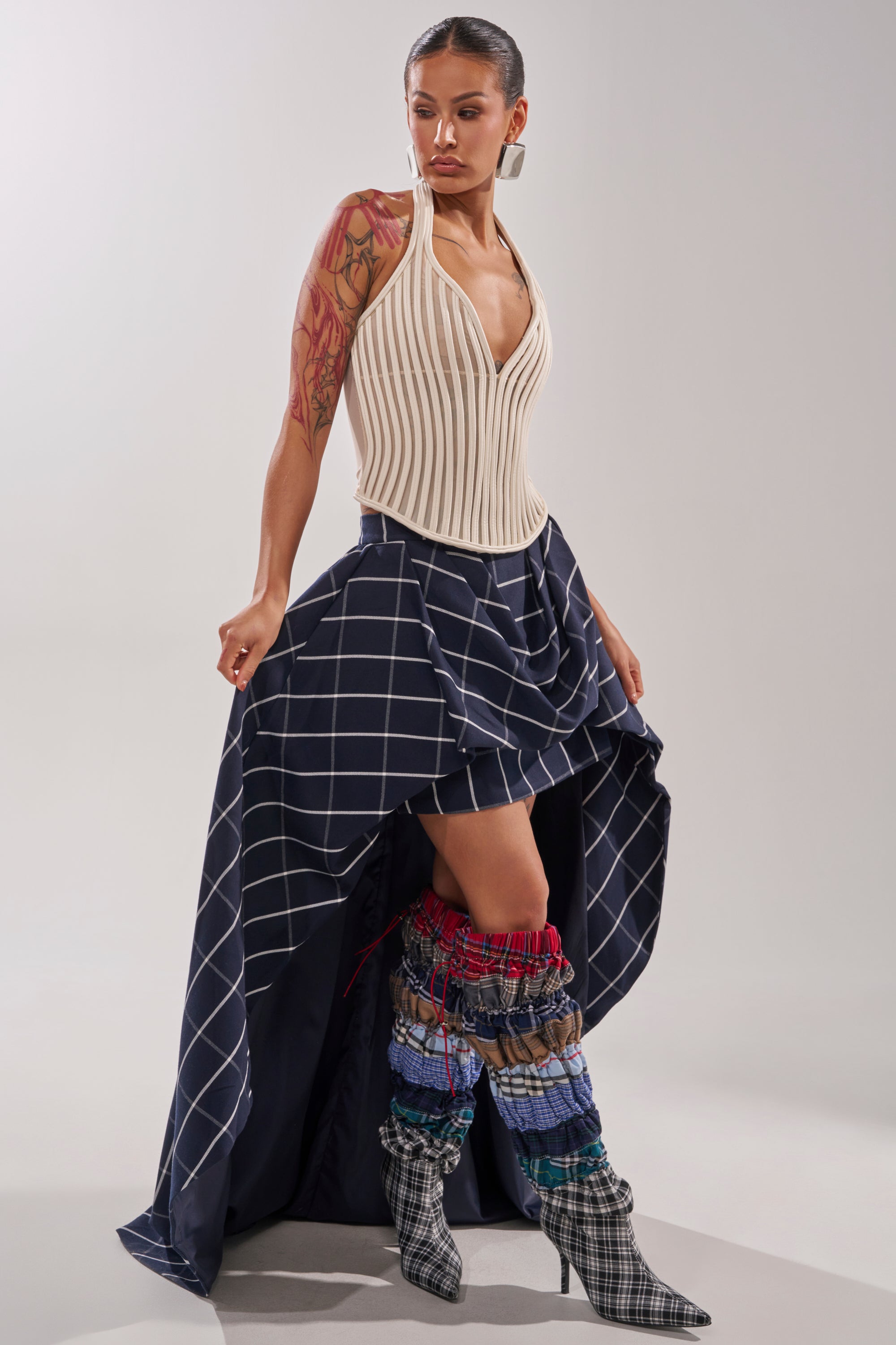 A woman poses in the MAKE A STATEMENT HIGH LOW SKIRT—a navy skirt with white grid lines—paired with a sleeveless cream corset top, tall patchwork boots, hoop earrings, and a sleek pulled-back hairstyle that reveals her large arm tattoo. 7