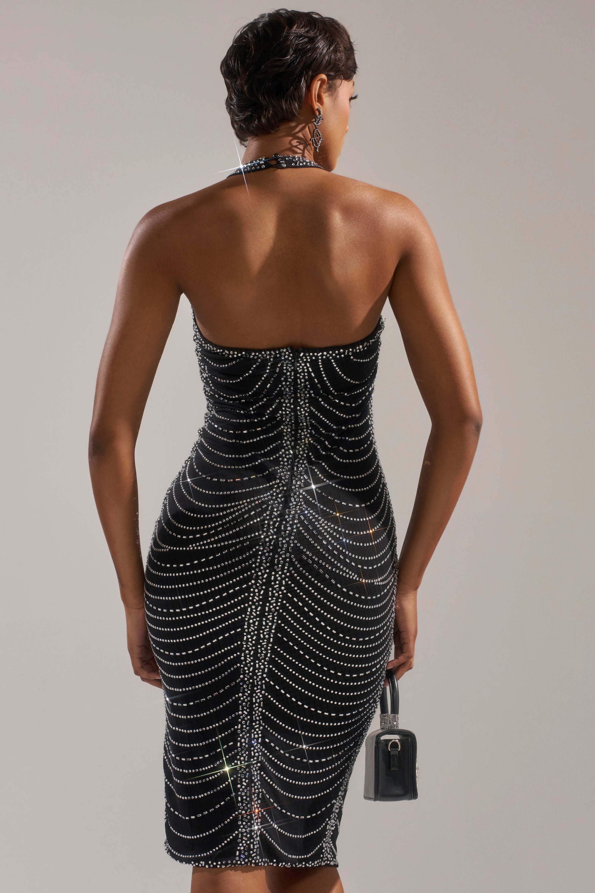 A person with short hair, seen from behind, wears the LANA RHINESTONE MIDI DRESS with sparkling lines and holds a small black handbag in their left hand against a neutral background.