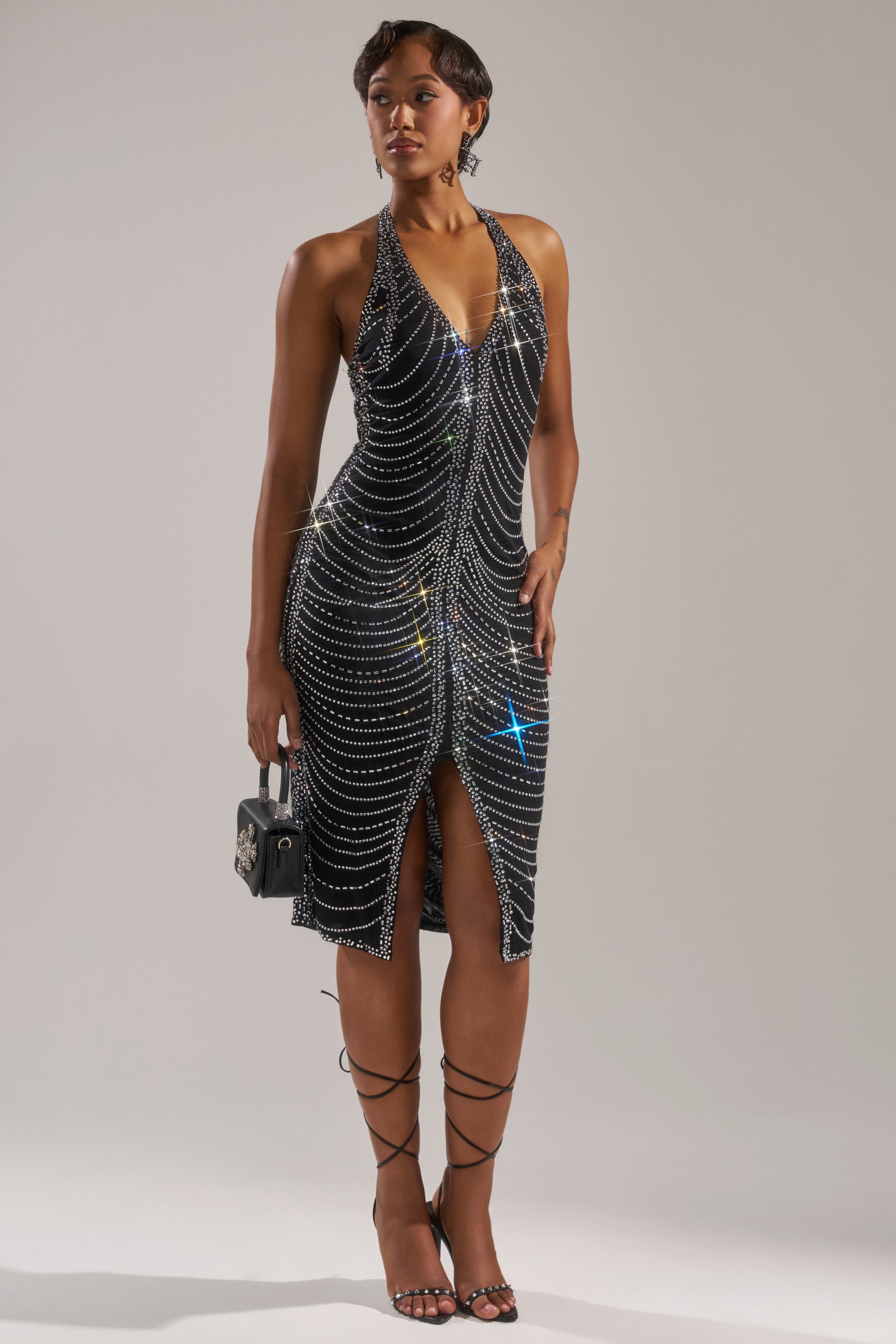Wearing the LANA RHINESTONE MIDI DRESS featuring a sleeveless, deep V-neck and glittering rhinestones for an hourglass effect, a woman pairs it with strappy heels and a small black handbag against a plain background.