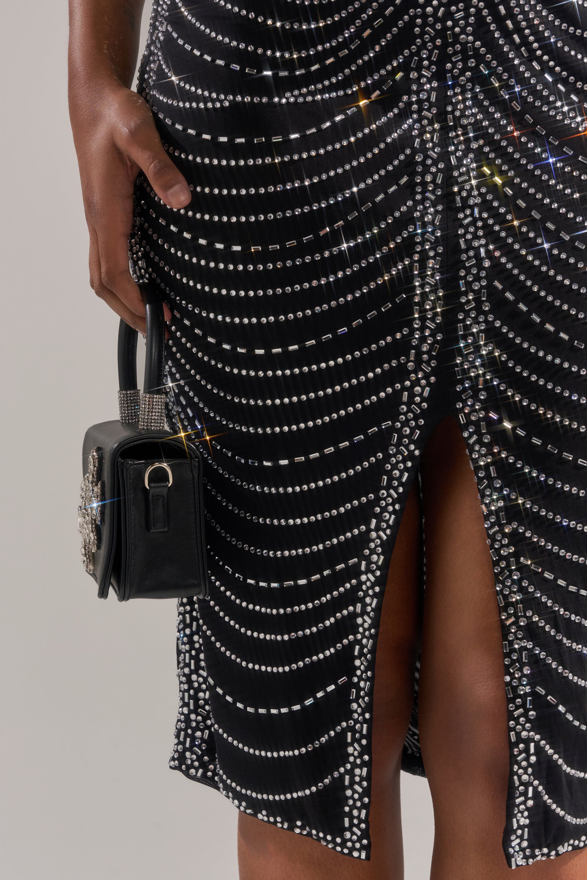 Wearing the LANA RHINESTONE MIDI DRESS embellished with sparkling stones, a person holds a small black bag with a rhinestone handle, standing with one hand by their side.
