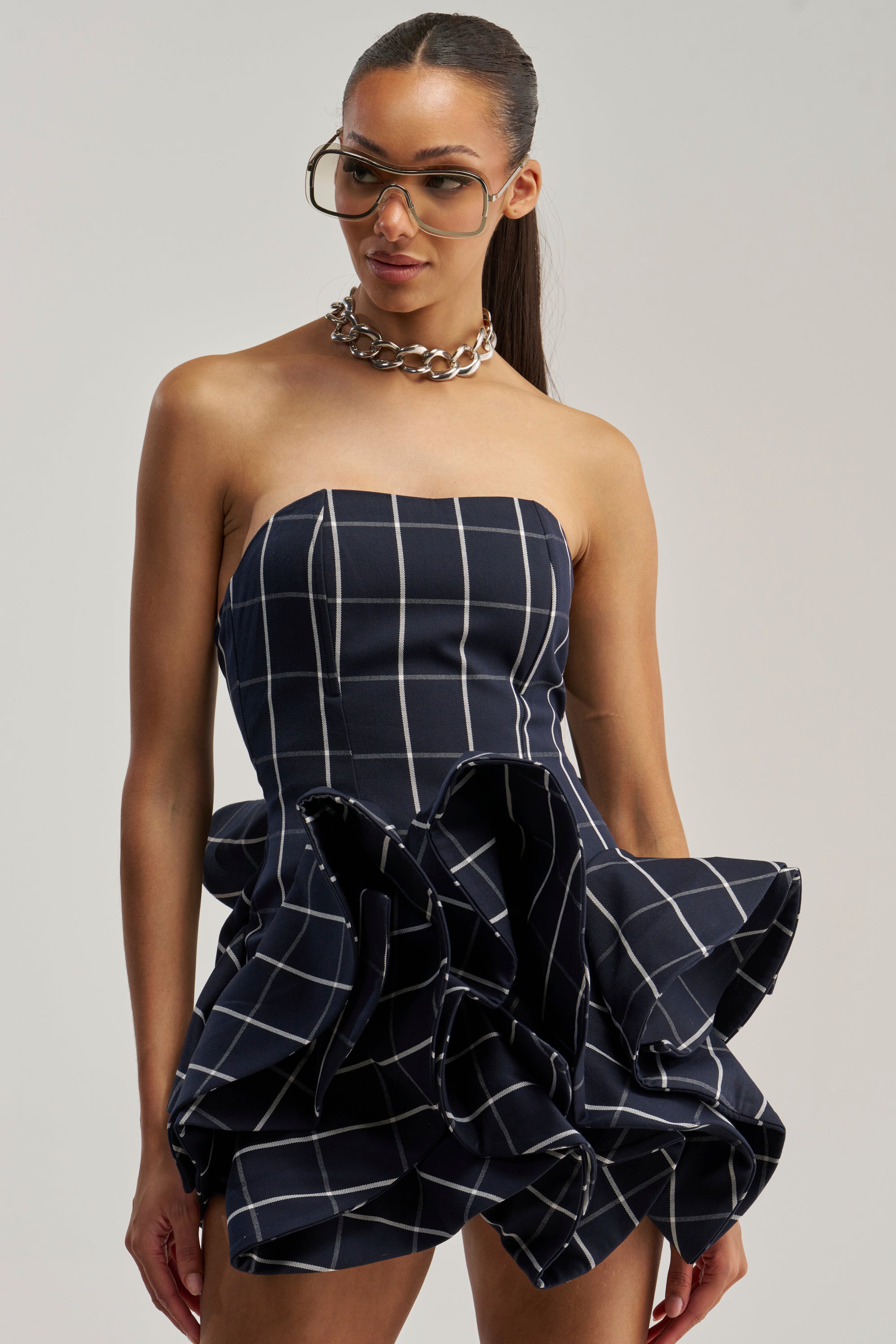 A woman stands against a plain light background wearing the SWEET ILLUSION BUBBLE SKIRT MINI DRESS—a strapless navy plaid mini dress featuring a corset-inspired bodice and ruffled skirt—paired with a chunky silver chain necklace and large glasses.