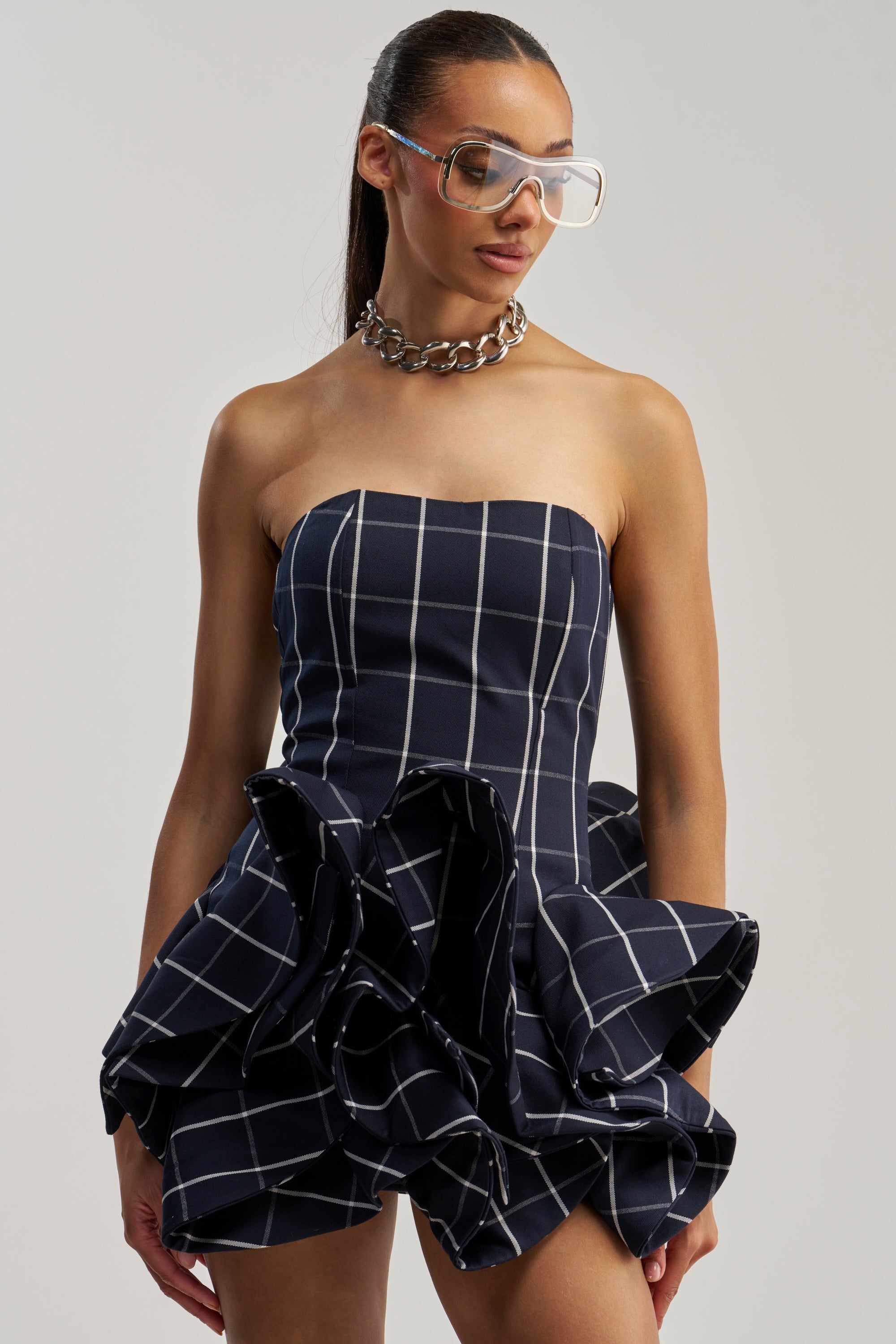 A woman wears the SWEET ILLUSION BUBBLE SKIRT MINI DRESS—a strapless navy mini dress featuring a white grid pattern and ruffled bubble skirt—paired with a chunky chain necklace, clear oversized glasses, and her hair pulled back.