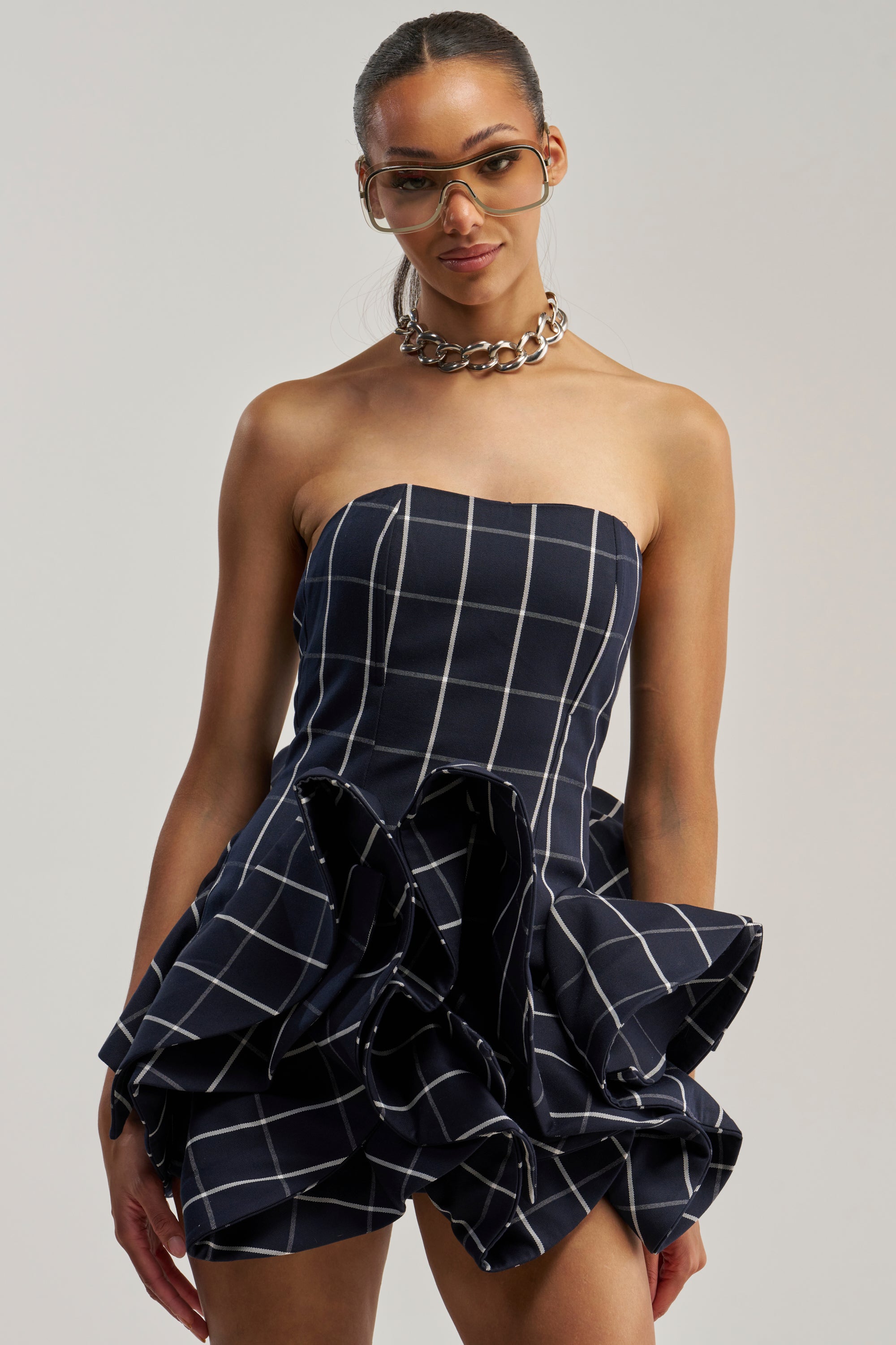 A woman wearing oversized glasses, a chunky chain necklace, and the SWEET ILLUSION BUBBLE SKIRT MINI DRESS—a strapless navy mini dress with white grid lines and a ruffled bubble hem—stands against a plain background.