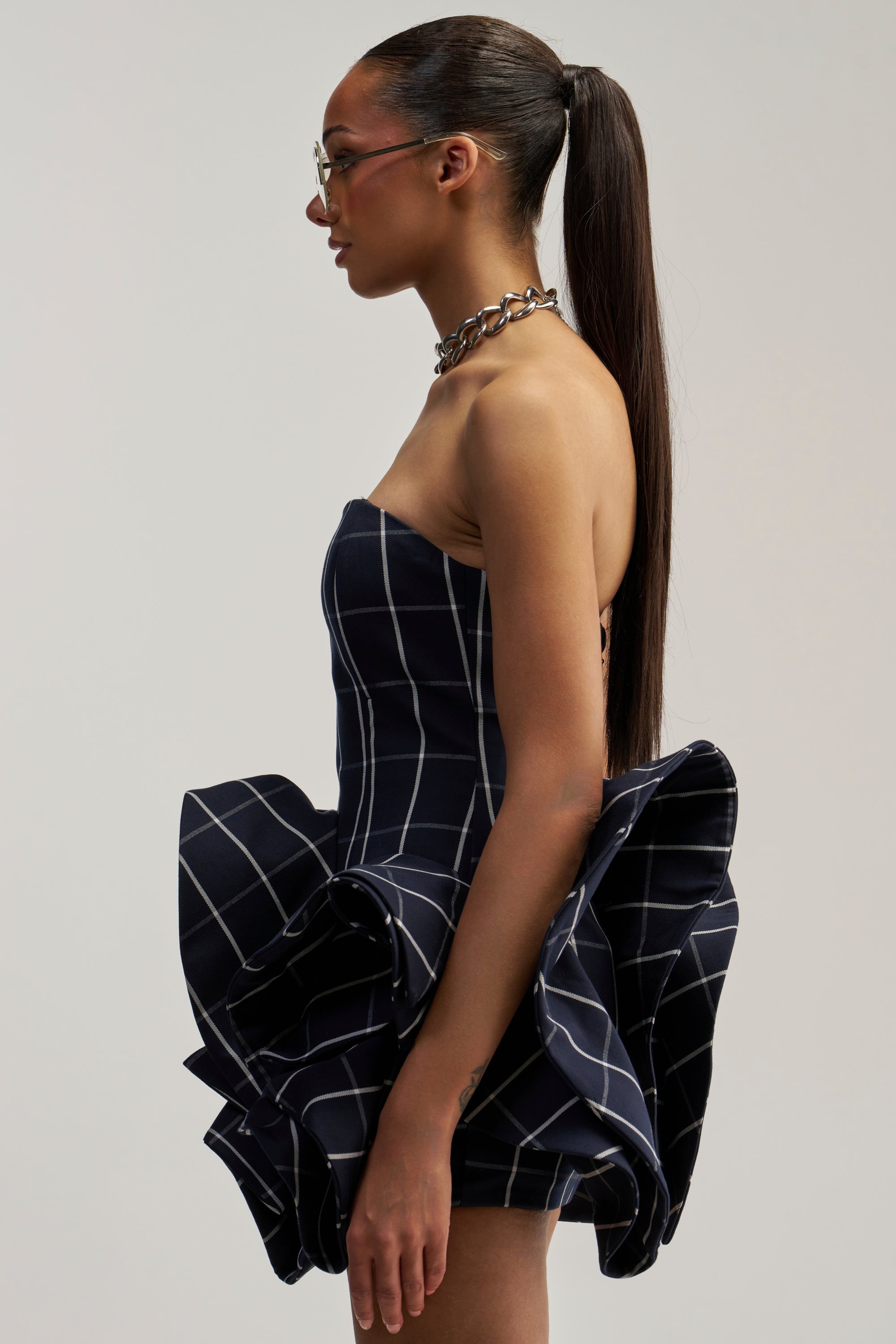 A woman in profile wears the SWEET ILLUSION BUBBLE SKIRT MINI DRESS, a black strapless piece featuring white grid lines and a dramatic ruffled skirt reminiscent of a bubble mini dress. She pairs it with a silver necklace and glasses, her long hair swept up in a high ponytail against a plain background.