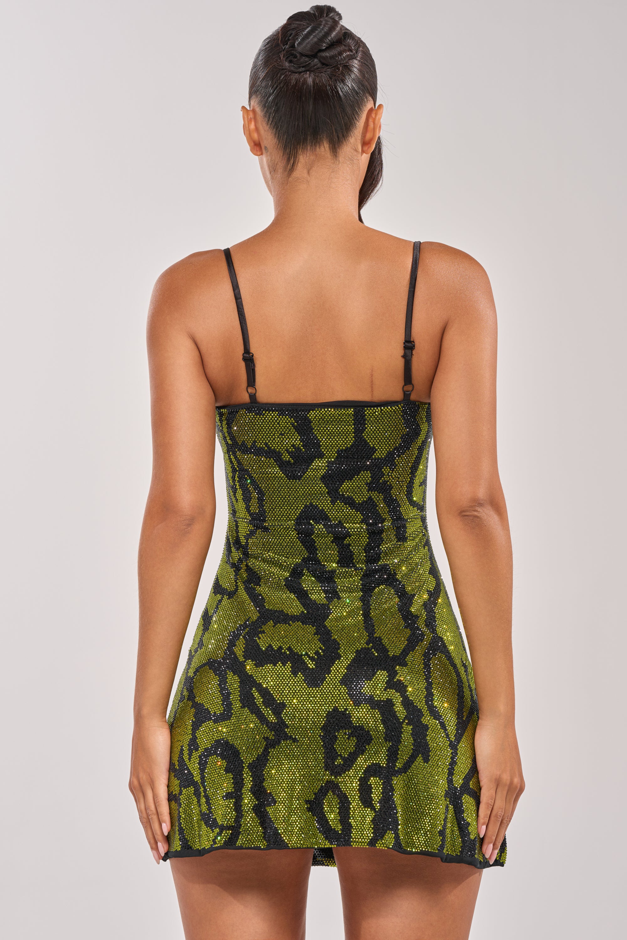 A woman with her hair in a high bun is shown from the back wearing the GABBI SEQUIN PRINT MINI DRESS IN GREEN, featuring a green and black leopard print with thin straps, standing against a plain light background. 4