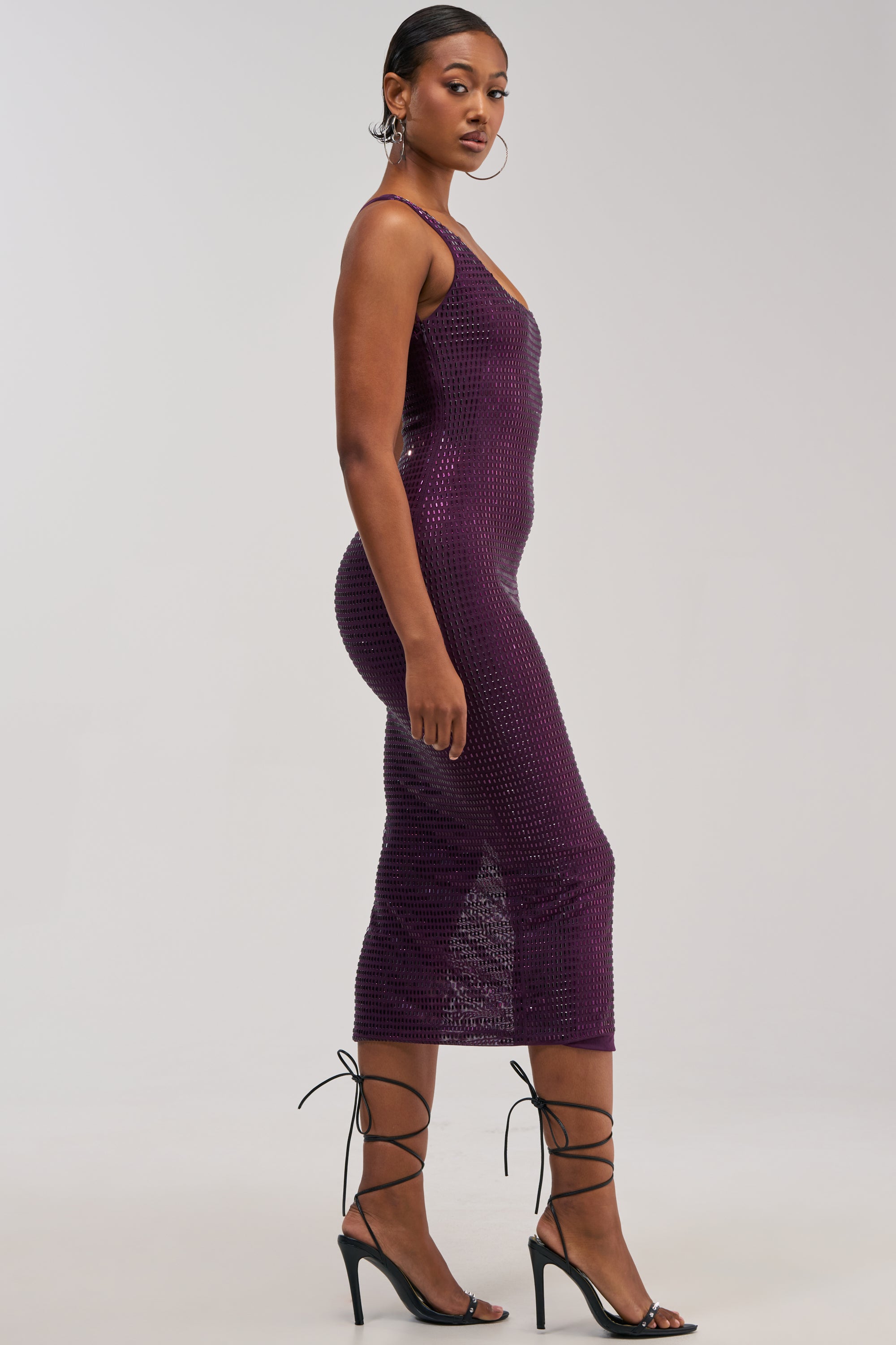 A woman stands in profile against a plain light background, wearing the MY MUSE EMBELLISHED RHINESTONE MIDI DRESS IN AMETHYST—a sleeveless, fitted, purple sequin dress adorned with rhinestone embellishments—paired with black strappy high heels.
