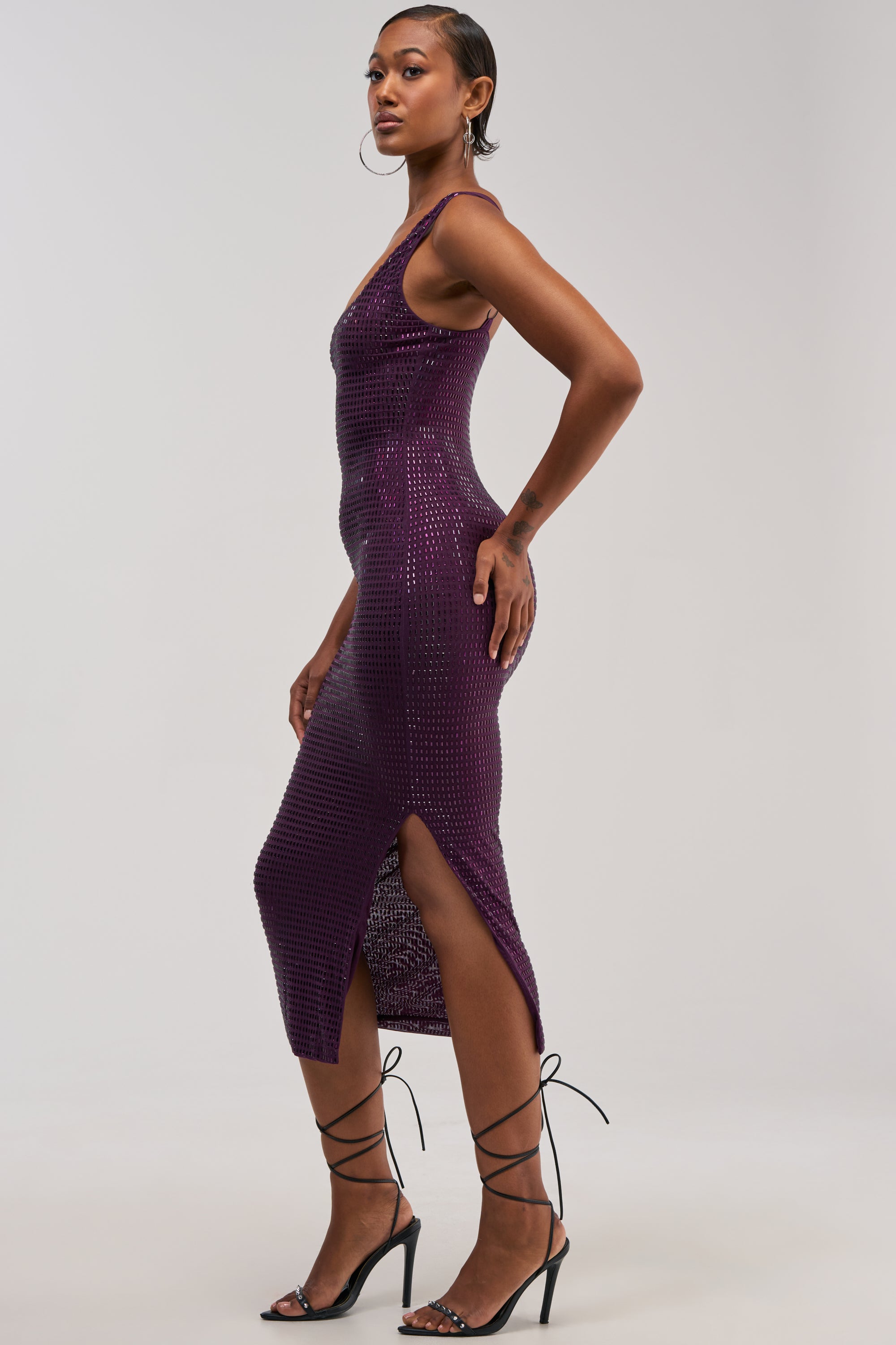 A woman poses sideways in the MY MUSE Embellished Rhinestone Midi Dress in Amethyst—a sleeveless, purple mesh dress adorned with sequins and rhinestones, featuring a side slit—paired with black strappy heels and hoop earrings against a plain background, making it perfect for special occasions.