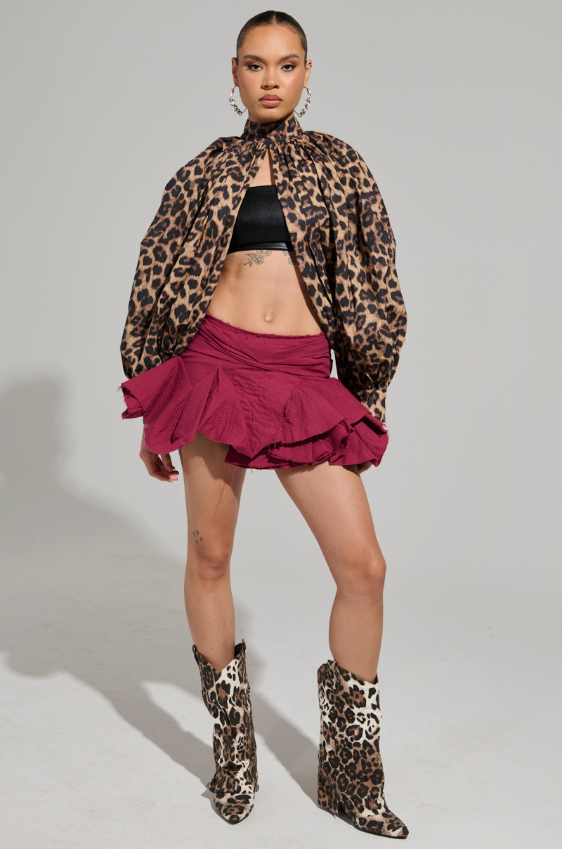 A person poses confidently with a leopard-print cape and matching boots, wearing a black crop top and the JANEY MICRO MINI RUFFLE SKIRT in vibrant fuchsia. They stand against a plain gray background.