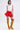 A woman stands against a plain background wearing a white sleeveless top with colorful circular details, the JANEY MICRO MINI RUFFLE SKIRT IN RED, and tall mustard-yellow lace-up high heel boots.