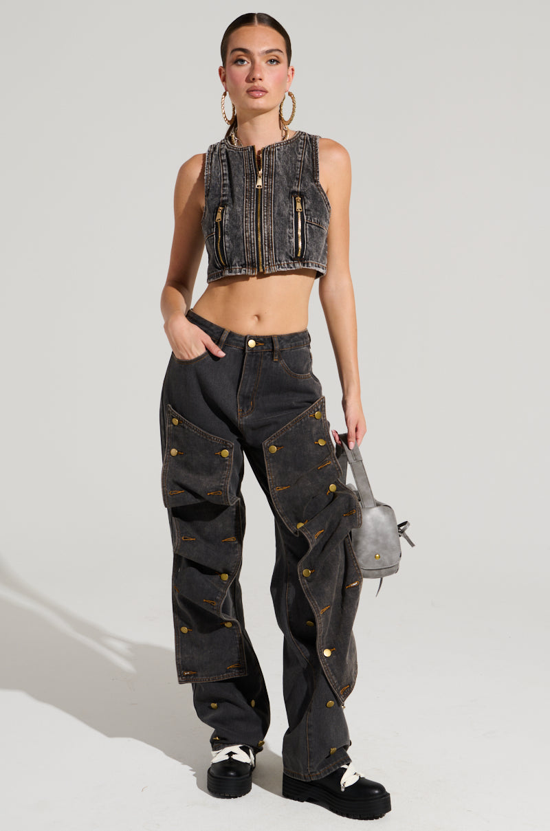In a studio setting, an individual models a dark denim crop top paired with the high-rise UNBUTTON ME DENIM JEANS, featuring a straight leg silhouette adorned with gold buttons. They accessorize with a small gray bag and wear black and white platform shoes, all set against a simple, light-colored backdrop.