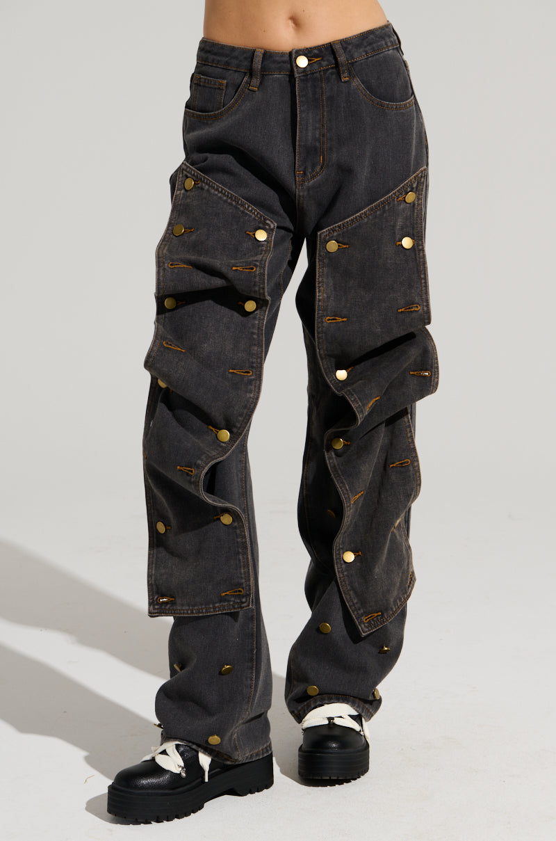 A person wearing the UNBUTTON ME DENIM JEANS, featuring a high rise with gold buttons and ruffled embellishments, stands on a light gray background. The jeans are paired with black shoes that have white laces.