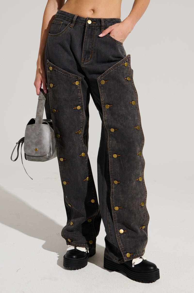 A person stands against a plain light background wearing the UNBUTTON ME DENIM JEANS, characterized by their high-waisted, wide-leg design and distinctive circular brass accents along the sides. They pair this sleek look with a small gray handbag and black platform shoes to create a striking silhouette.