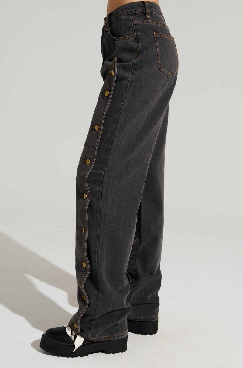 Side view of a person wearing UNBUTTON ME DENIM JEANS in dark gray, showcasing a button-up seam along the side. The high-rise denim features a straight leg silhouette, perfectly paired with black shoes. The background is plain and light-colored.