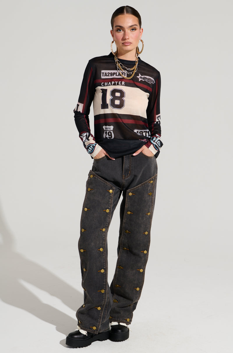 Standing confidently, a person wears a black and white long-sleeve shirt adorned with graphic designs and numbers. Their hands rest in the pockets of high-rise UNBUTTON ME DENIM JEANS featuring a wide-leg silhouette. Chunky black shoes enhance their ensemble, accompanied by large hoop earrings that elegantly frame their face.