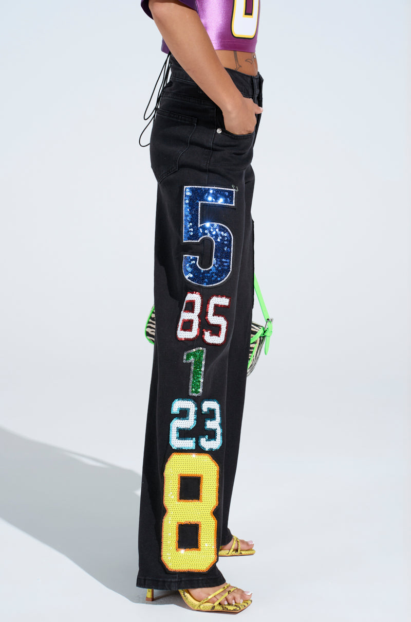 Against a light background, someone stands out in LUCKY NUMBER ONE DENIM PANT. These black pants feature oversized sequined numerical patches. Paired with a purple top, yellow heels, and a green bag, the ensemble delivers a striking mix of bold colors and playful fashion elements.