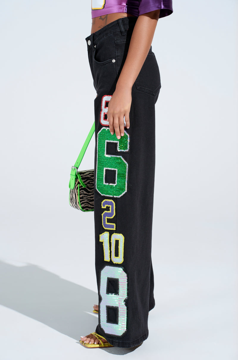 Against a light background, an individual sports LUCKY NUMBER ONE DENIM PANT with vibrant numerical patches. They hold a small zebra-patterned bag with a lime green handle and wear a purple top, proudly embracing the bold style of wide-leg pants.