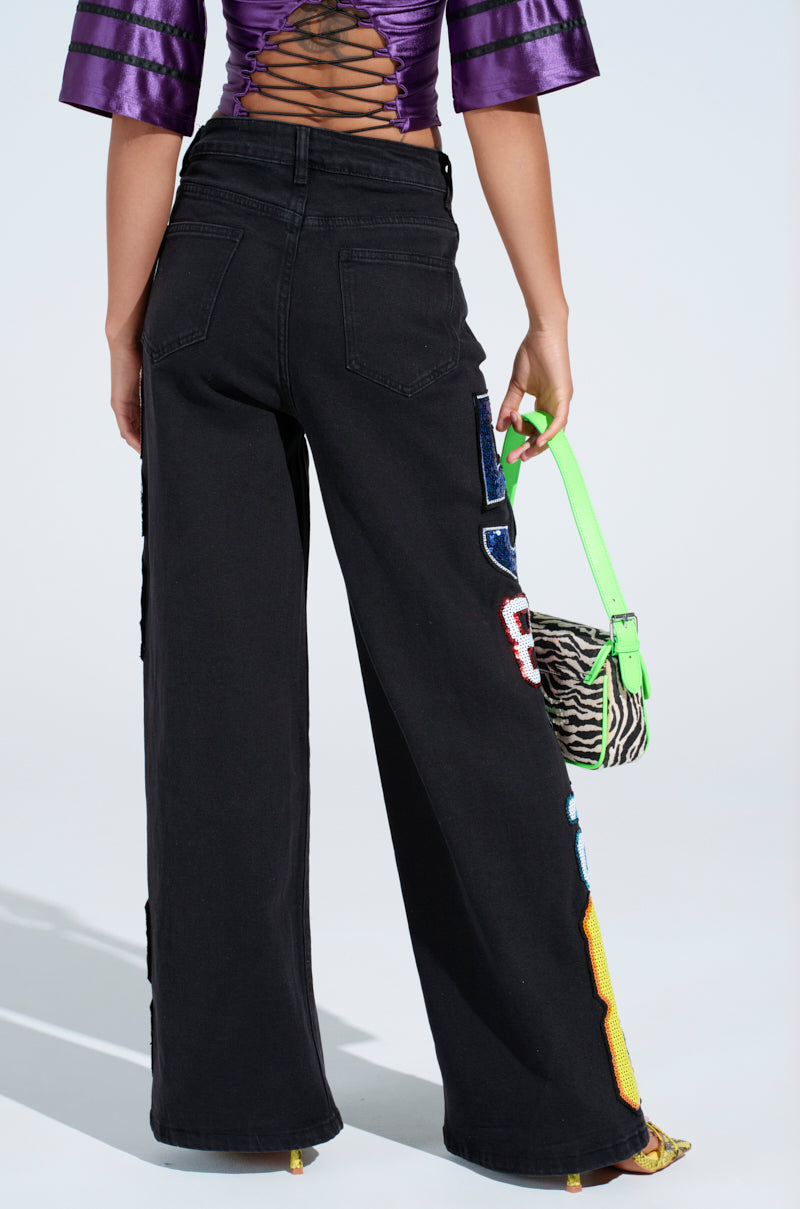 Wearing the LUCKY NUMBER ONE DENIM PANT in black with colorful numerical patches, paired with a purple lace-up back top and holding a green zebra-patterned handbag, the look is stylishly modern with chic wide-leg denim and vibrant patterns.