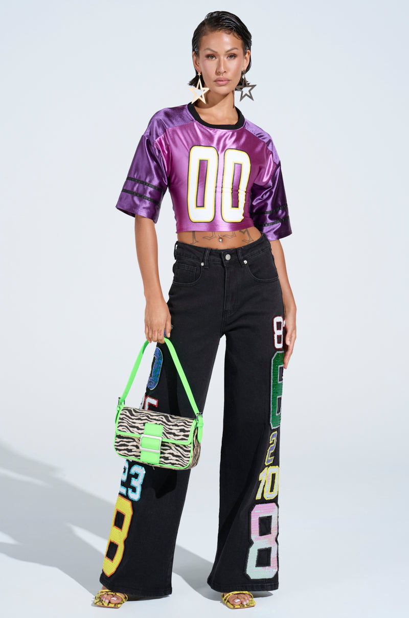 A person wears a shiny purple crop top with "00" and the LUCKY NUMBER ONE DENIM PANT featuring colorful numerical patches. Yellow heeled sandals add flair, while a zebra-patterned handbag with a green strap complements the look. Large star-shaped earrings complete this stylish ensemble.