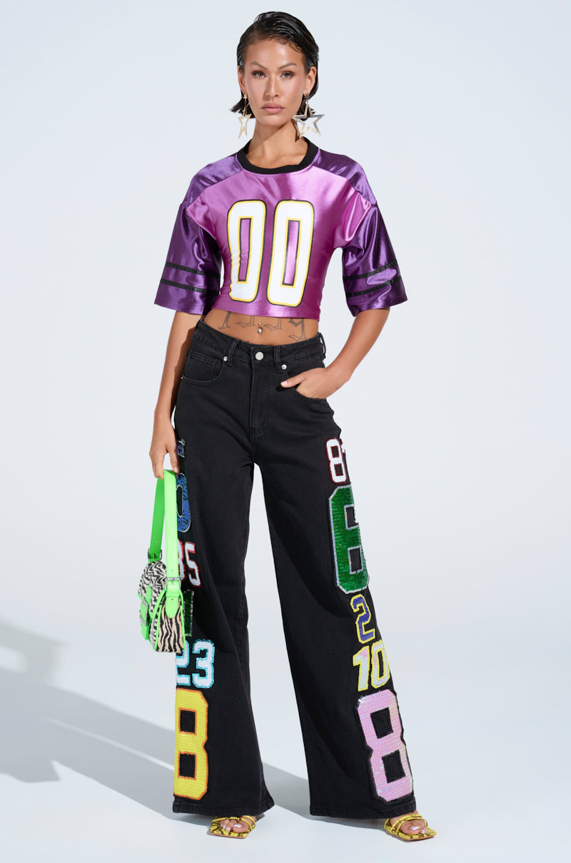 A person poses confidently in a purple crop top with "00" on the front and black wide-leg LUCKY NUMBER ONE DENIM PANT adorned with numerical patches. They hold a green and black tote bag and wear yellow sandals, embracing a bold, stylish wide-leg silhouette.
