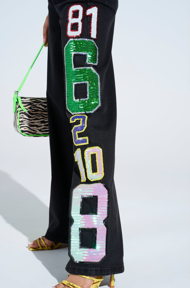 Close-up of a person wearing LUCKY NUMBER ONE DENIM PANT with sequined patches (81, 6, 2, 10, 8), a zebra-print bag with a bright green strap, and yellow strappy sandals.