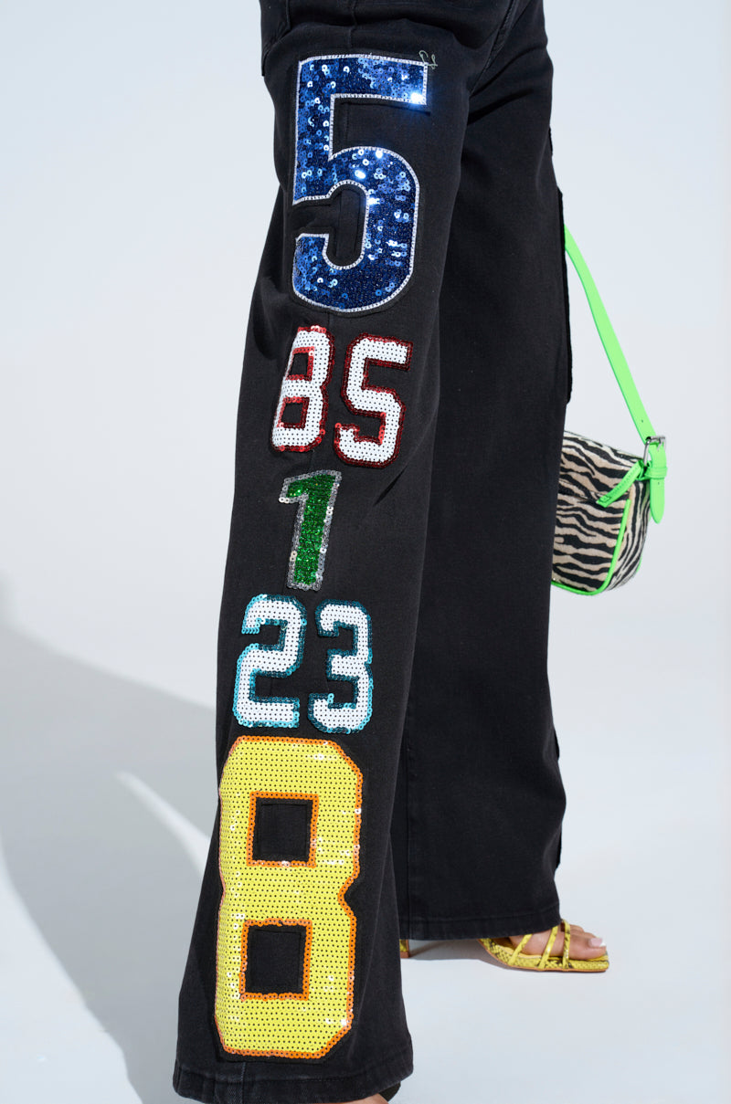 Close-up of LUCKY NUMBER ONE DENIM PANT featuring vibrant "5," "85," "1," "23," and "8" patches on the leg, paired with a zebra-patterned bag with a green strap and yellow heels. High-rise design adds style to this bold look.