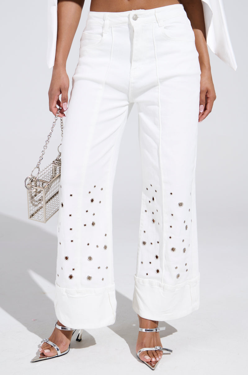 Someone is wearing JULIA CROPPED WIDE LEG JEANS with decorative holes and silver embellishments near the ankles, paired with a white top, silver high-heeled sandals, and a sparkly silver handbag against a plain white background.