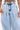 A person wears the IN CHARGE WIDE LEG DENIM PANT: high-rise, light-wash, wide-leg jeans with pleats. The cropped image shows from waist to mid-thigh, revealing part of their tattooed torso.