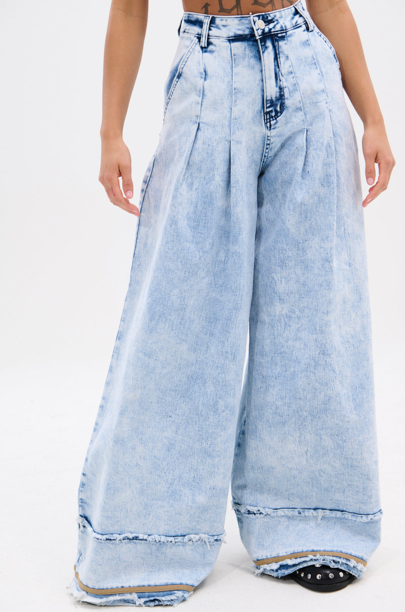 A person shows off the IN CHARGE WIDE LEG DENIM PANT—vintage-inspired, light-wash jeans with a high-rise waist, pleats, wide legs, and frayed hems—against a plain white backdrop. Only the lower body is visible.