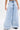 A person shows off the IN CHARGE WIDE LEG DENIM PANT—vintage-inspired, light-wash jeans with a high-rise waist, pleats, wide legs, and frayed hems—against a plain white backdrop. Only the lower body is visible.