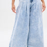 A person shows off the IN CHARGE WIDE LEG DENIM PANT—vintage-inspired, light-wash jeans with a high-rise waist, pleats, wide legs, and frayed hems—against a plain white backdrop. Only the lower body is visible.