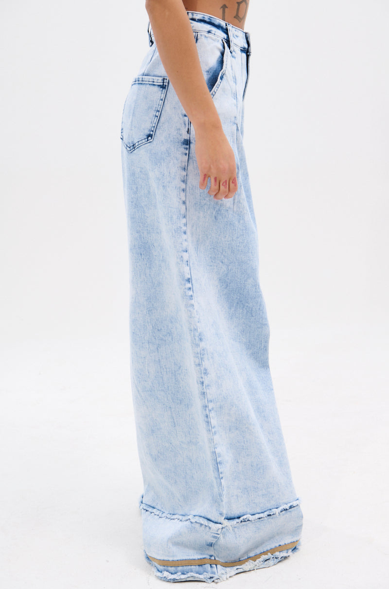 The lower half of a person stands sideways against a plain white background, wearing the IN CHARGE WIDE LEG DENIM PANT with a high-rise waist and raw hems for a vintage-inspired look.