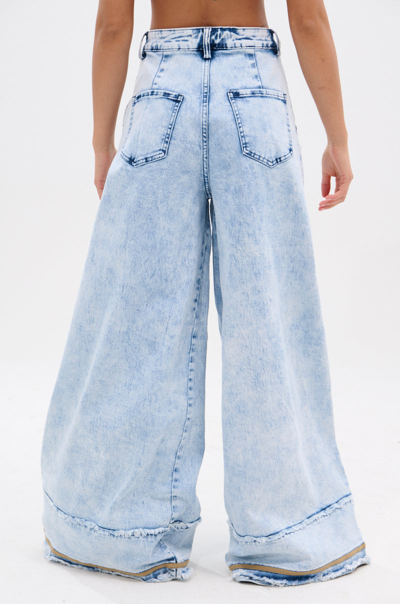 A person wearing the IN CHARGE WIDE LEG DENIM PANT, featuring a high-rise waist, light-wash, wide leg fit with large back pockets and frayed hems, stands with their back to the camera against a plain white background.